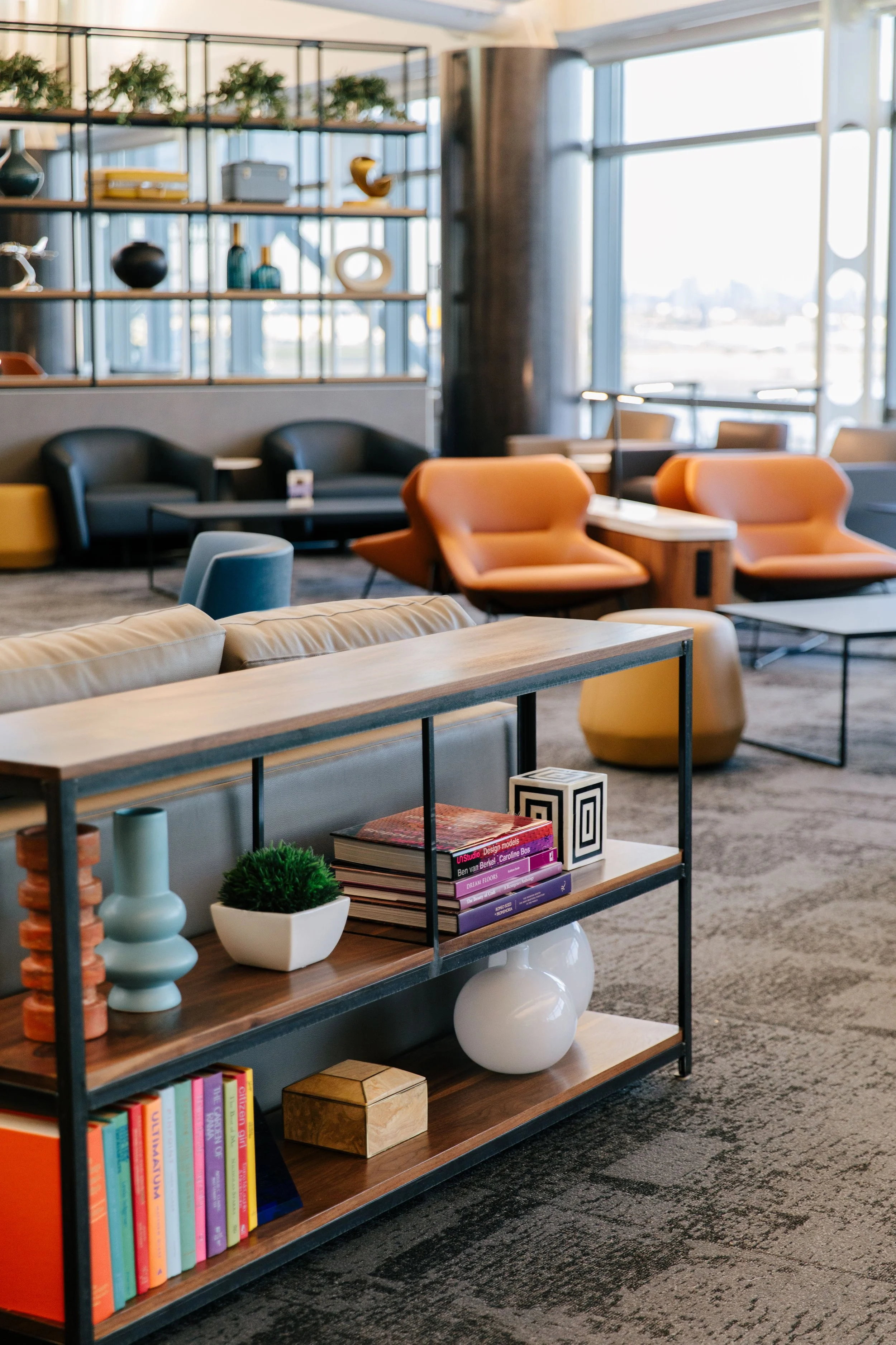 Modern lounge area with colorful chairs, a bookshelf with decorative items and books, and large windows letting in natural light.