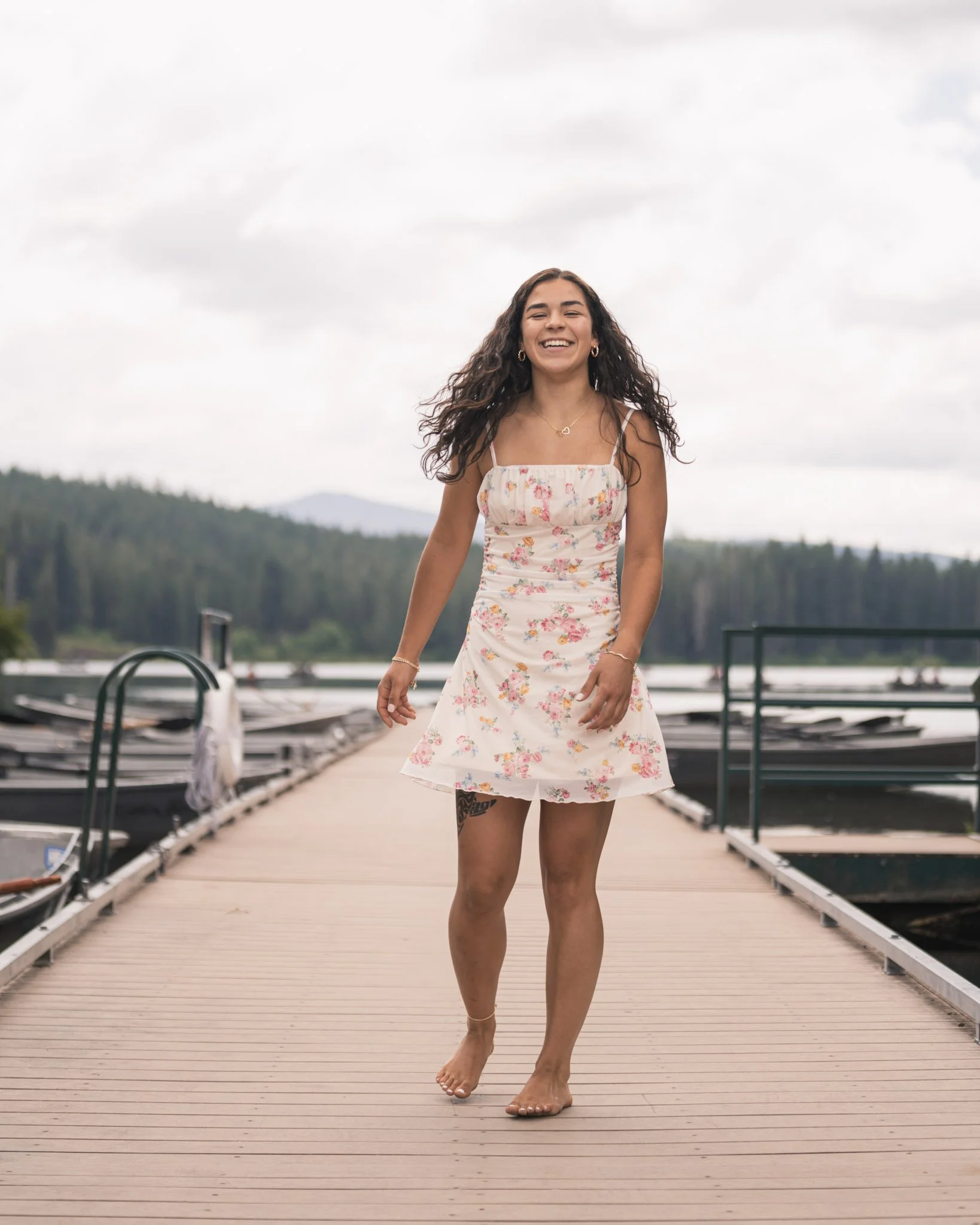 A young woman in a floral dress walking barefoot on a wooden dock beside a lake, with forested hills and cloudy sky in the background.
