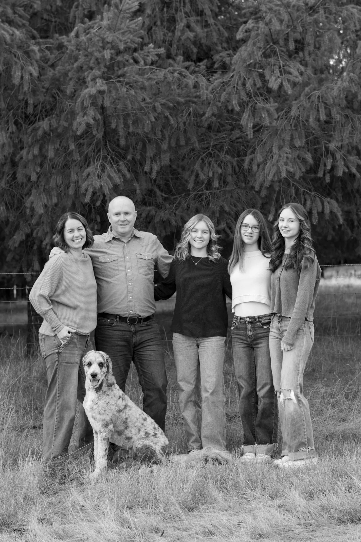 A family of five with a dog standing outdoors on grass in front of a large pine tree, smiling at the camera in a black and white photo.