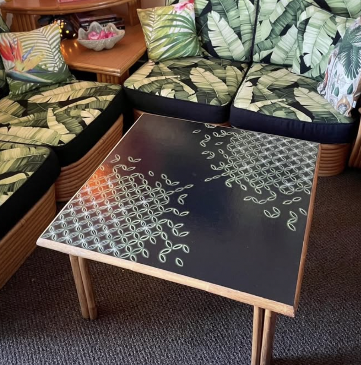 A rectangular table with a black top and a white geometric pattern, surrounded by couches with tropical leaf-patterned cushions in a living room.