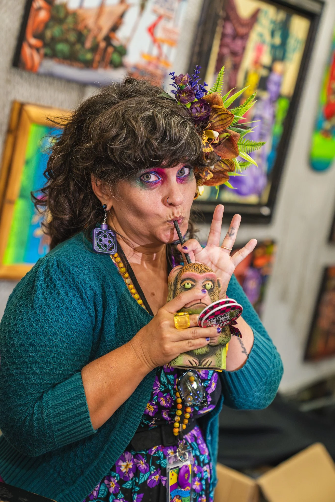 A woman with curly hair wearing vibrant makeup, a floral headpiece, and colorful accessories, is sipping from a cup with a straw. She is holding a monkey-shaped figurine and is standing in an art gallery with colorful artwork in the background.