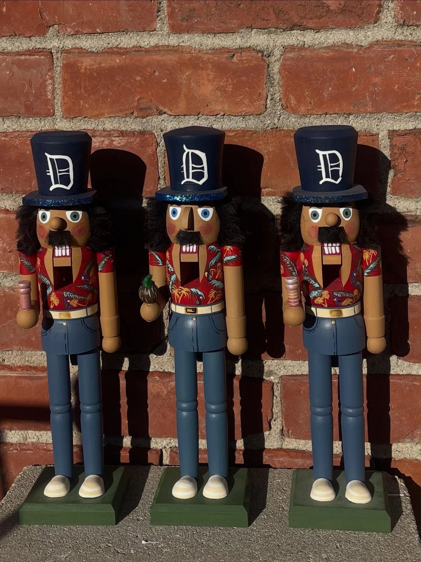 Three decorative nutcracker figures standing against a brick wall, each wearing a navy blue top hat with a white letter D, red shirts with tropical patterns, and blue pants, with long black hair and a mustache.