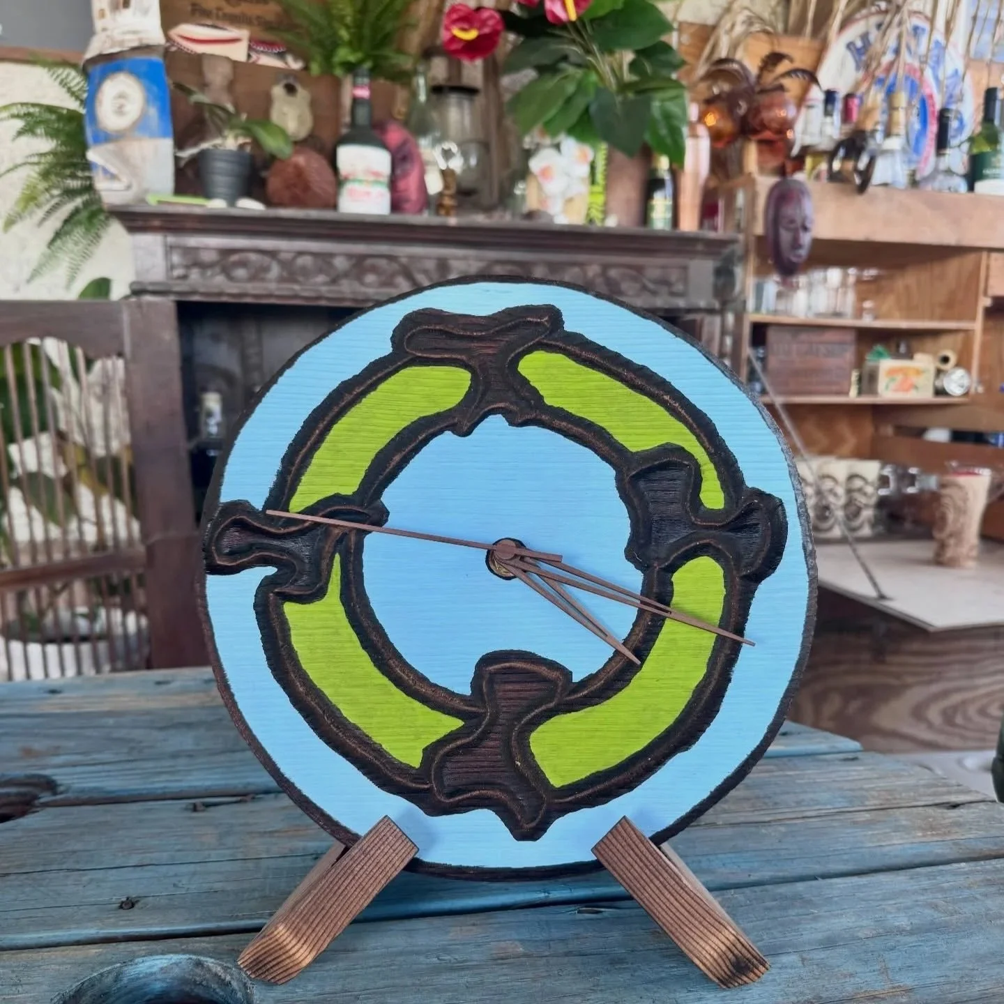 Handmade clock with a design resembling a reptile, painted in green and black, on a wooden easel. The background features a rustic setting with plants, paintings, and decorative objects.