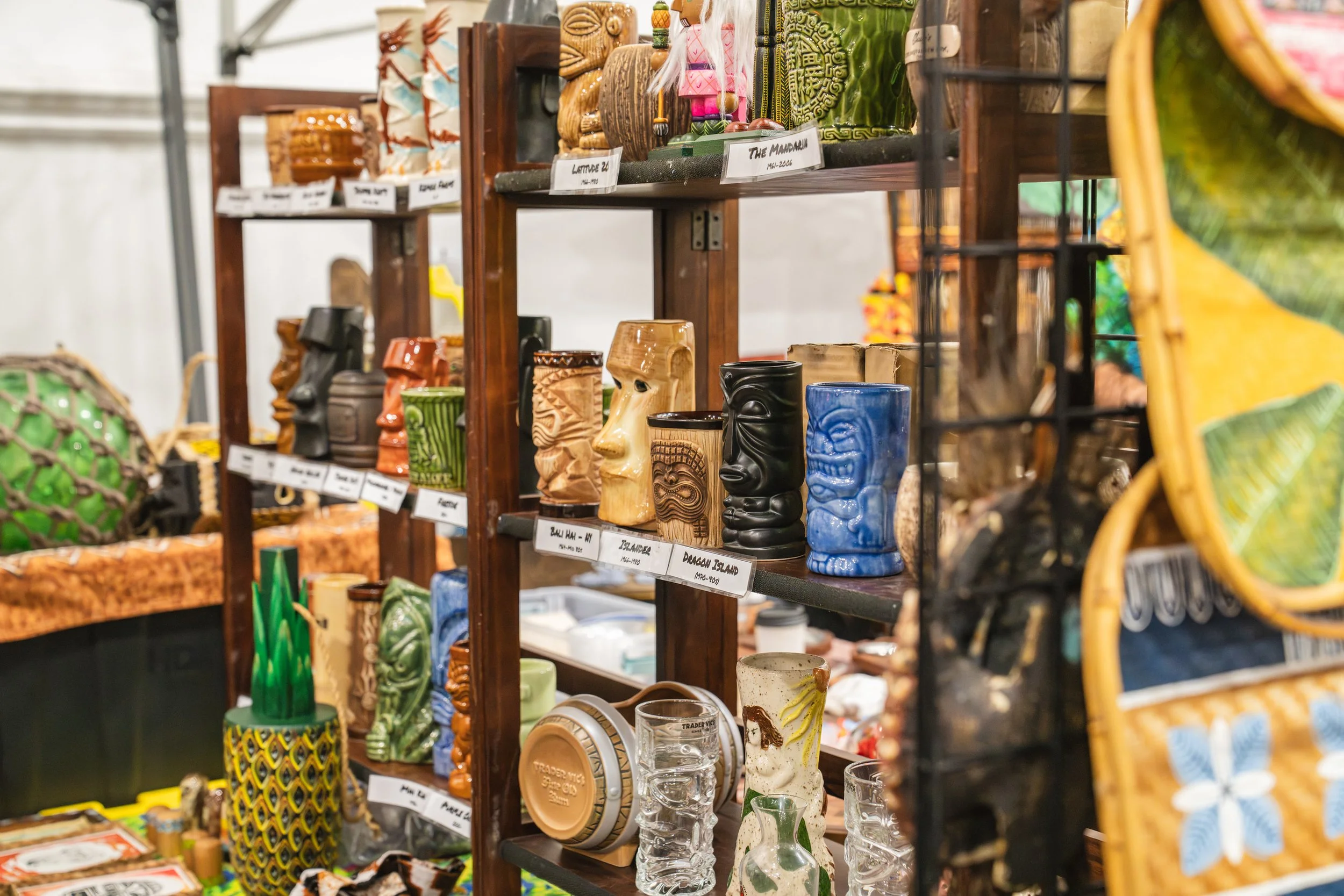 Display of various Polynesian and Hawaiian-style carved wooden tiki mugs and cups on wooden shelves at a market or fair.