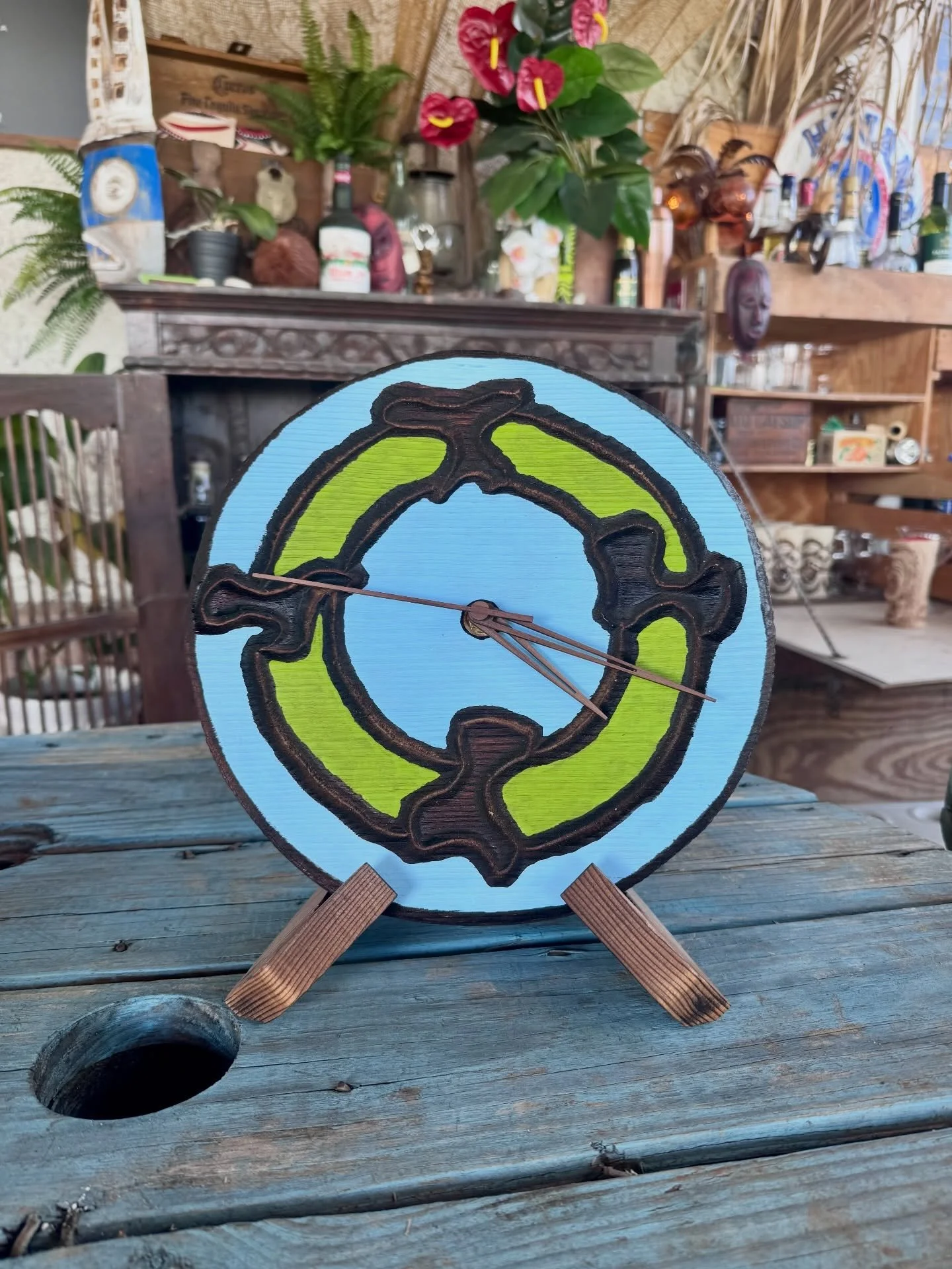 A painted round wooden clock with black and green design, standing on a small wooden easel on a wooden table, with a cluttered wooden shelf and plants in the background.
