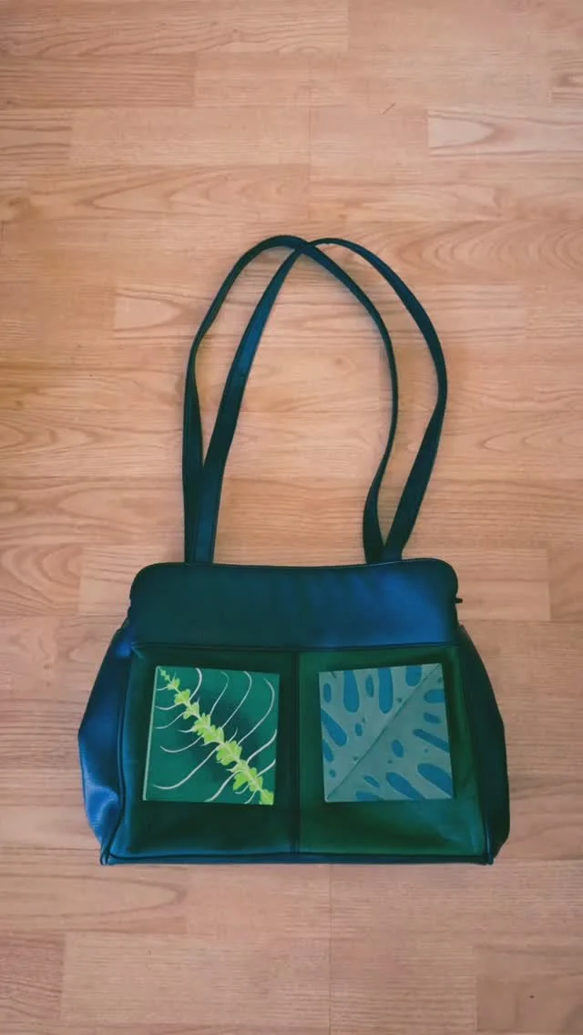 A black bag with two transparent pockets on the front, each containing a leaf print, and long black straps on a wooden floor.