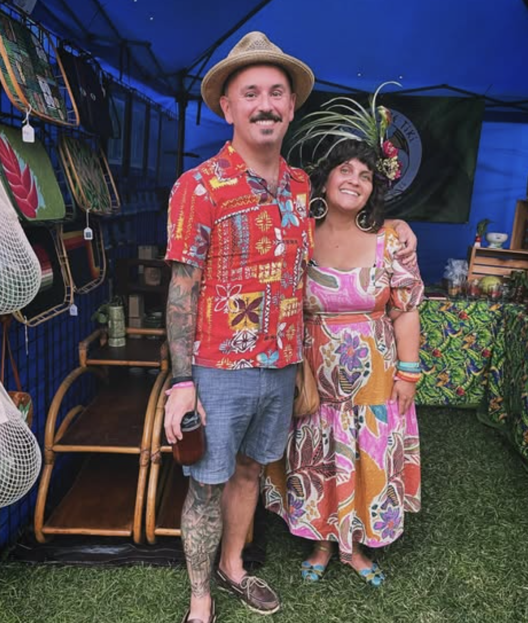 A man and woman smiling and posing inside a colorful booth at an outdoor event, with Hawaiian-style clothing and accessories, surrounded by leis and tropical decorations.