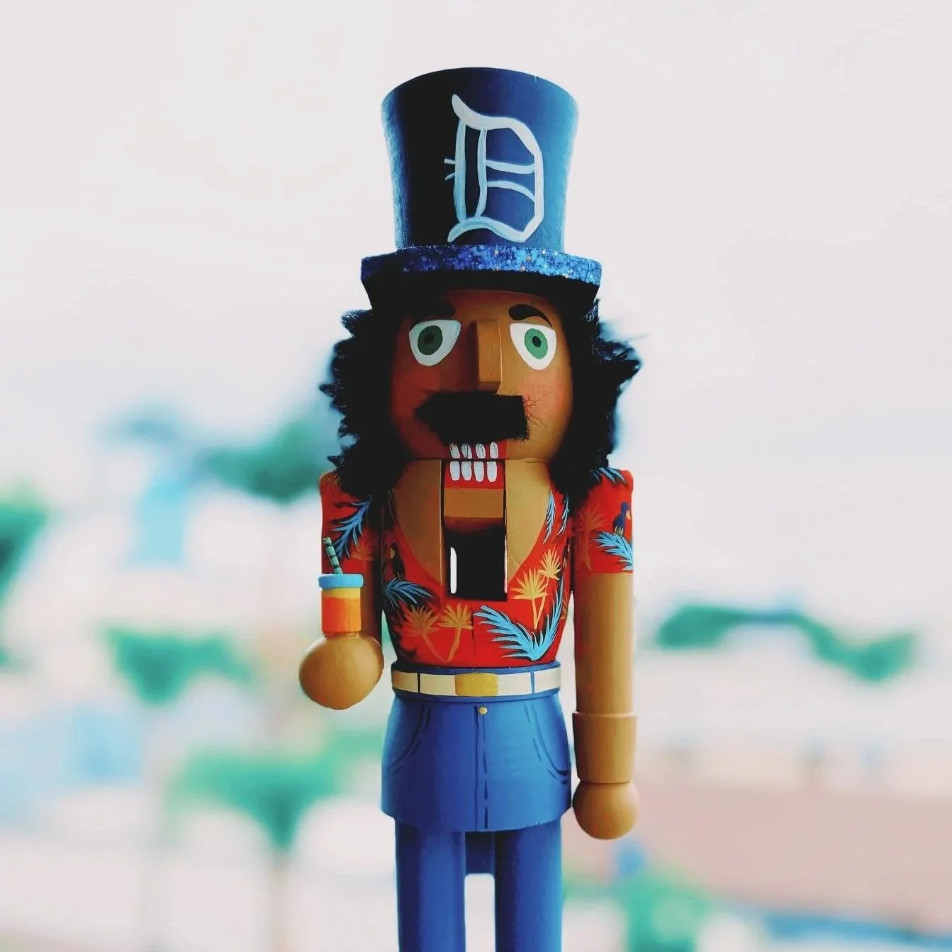 Wooden nutcracker figurine dressed as a man with a Hawaiian shirt, holding a drink, and wearing a tall top hat with a 'D' on it.