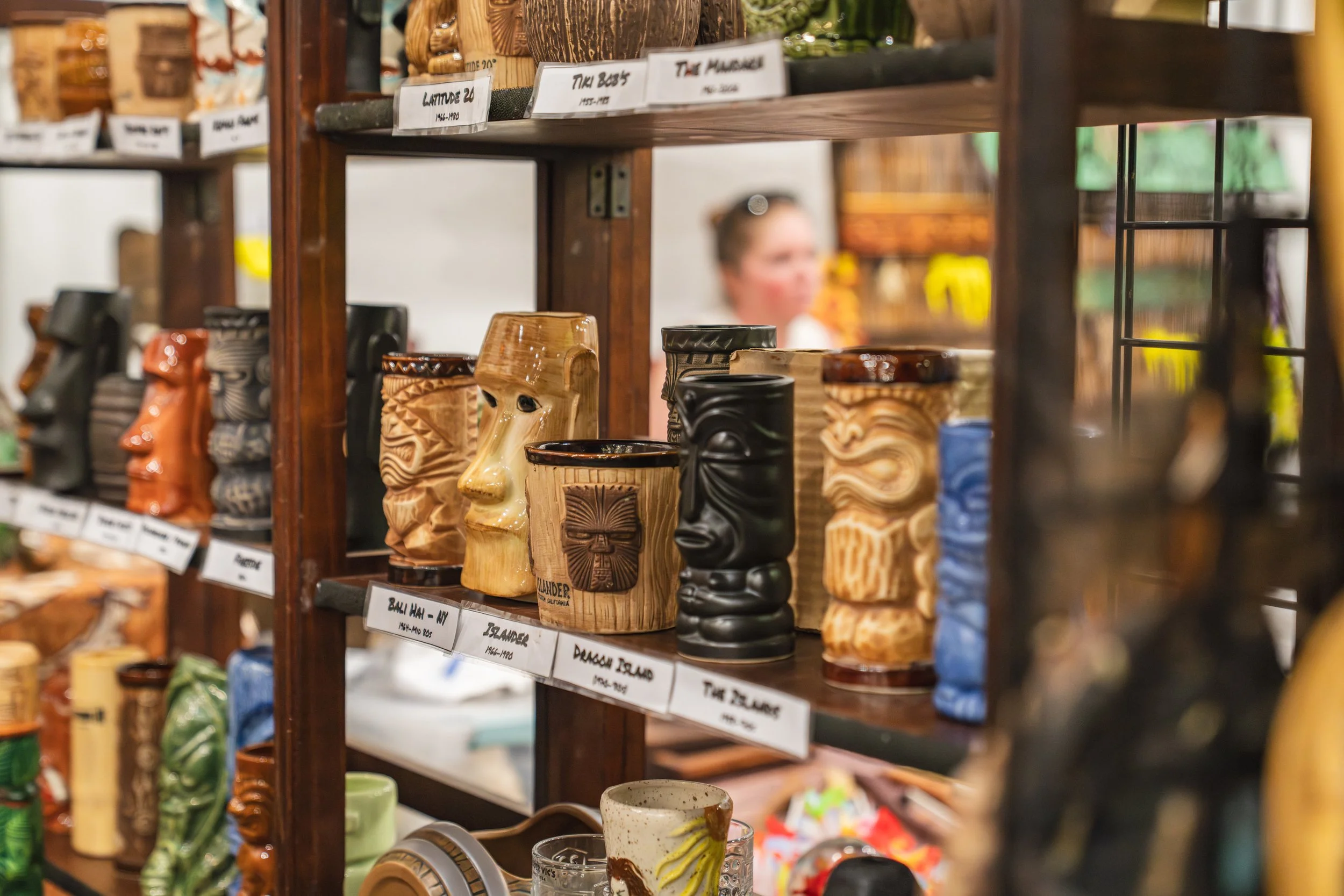 A display of tiki mugs on wooden shelves in a store, with some labels indicating locations such as the Flinders and Hawaii.
