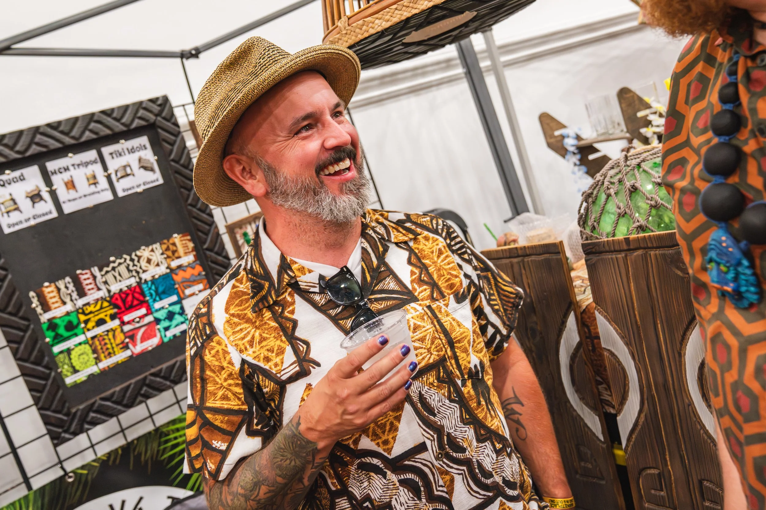 A smiling man with a gray beard, wearing a straw hat and a patterned tropical shirt, holding a clear plastic cup at an indoor market or craft fair. There are colorful woven items and signs with furniture images in the background.