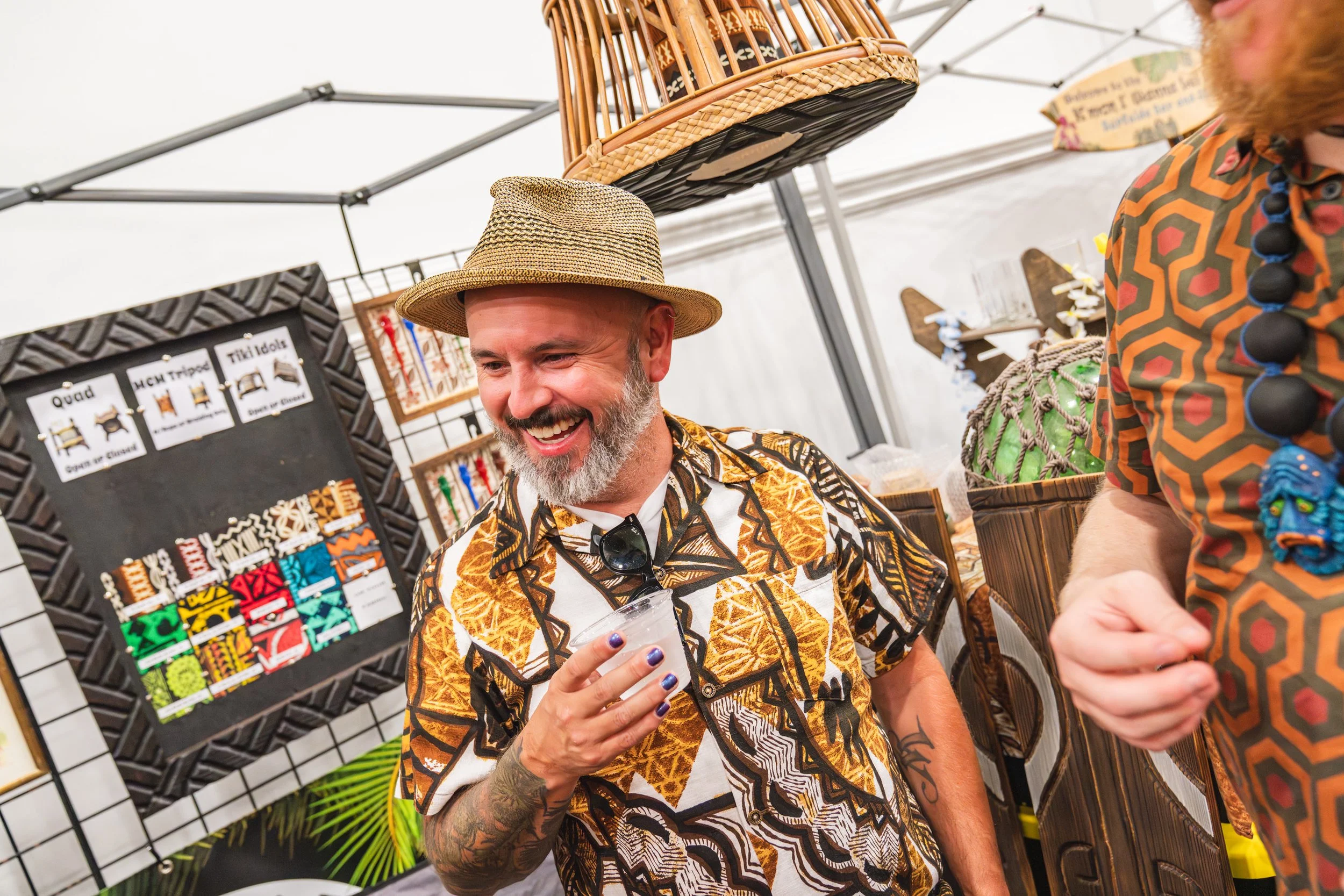 A man with a beard smiling, wearing a patterned shirt and a straw hat, is holding a drink and standing in front of a display of colorful woven items at an outdoor market.