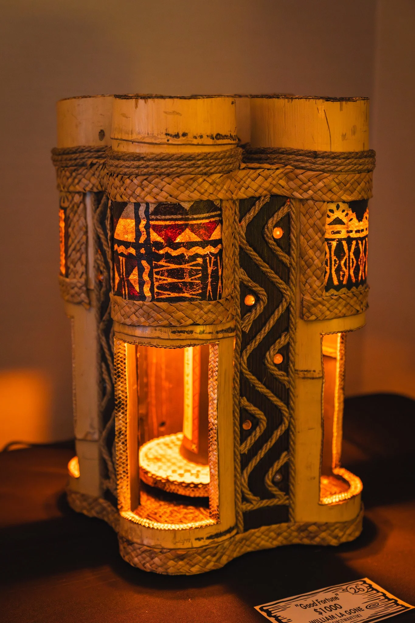Decorative lamp made of bamboo with painted tribal designs, illuminated from within, placed on a dark surface next to a $1000 'Good Fortune' lottery ticket.
