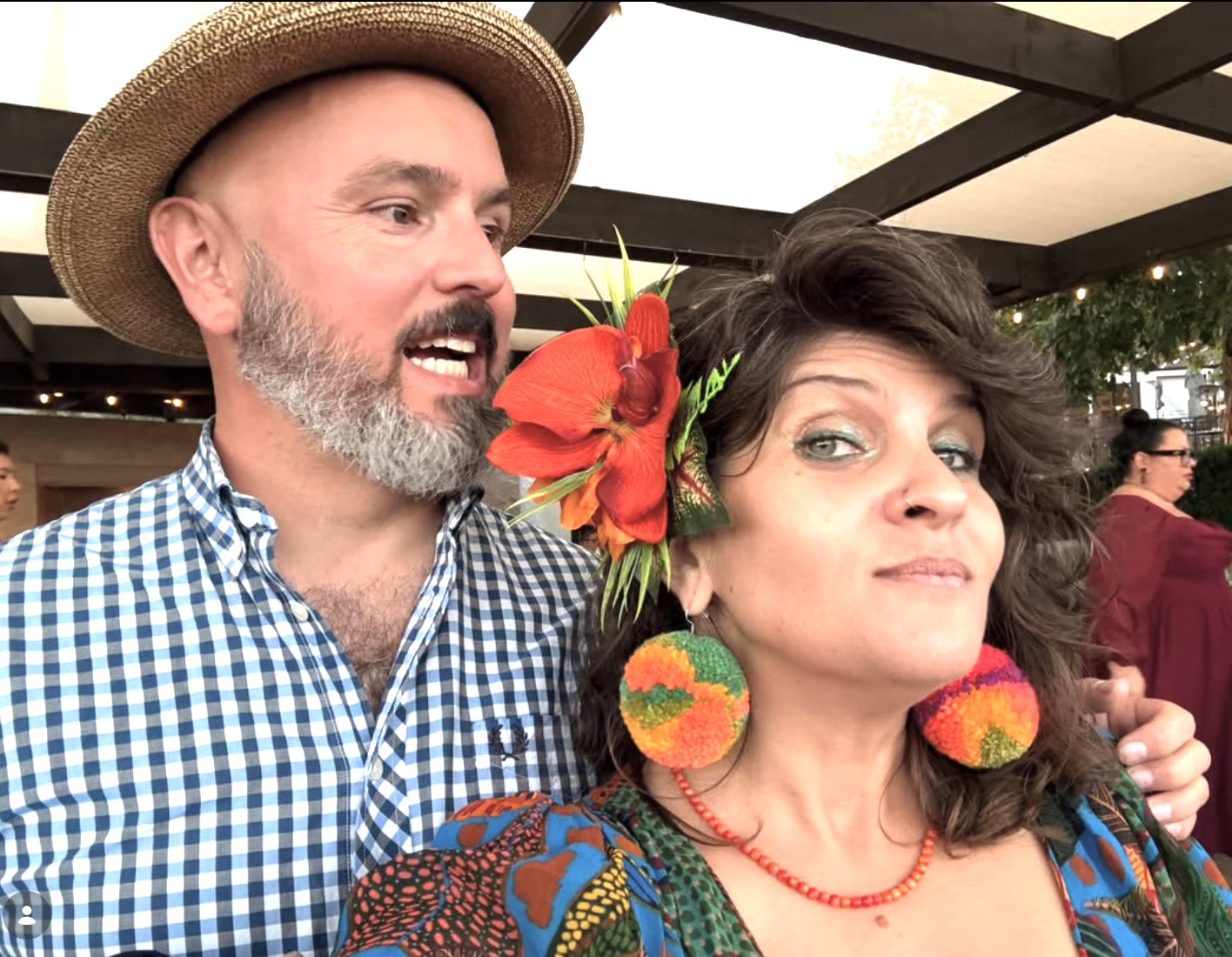 A man with a beard wearing a straw hat and a checkered shirt, standing next to a woman with wavy hair decorated with a large red and orange flower, colorful earrings, and a colorful dress. They appear to be at an outdoor event.