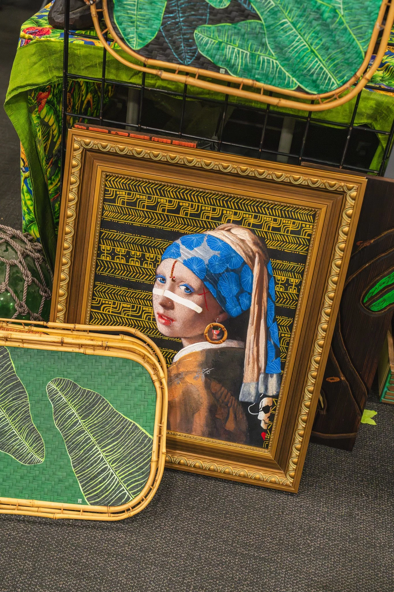 Display of framed art and decorative items on a patterned green and tropical-themed table cover with a black wire stand and a dark carpet floor.