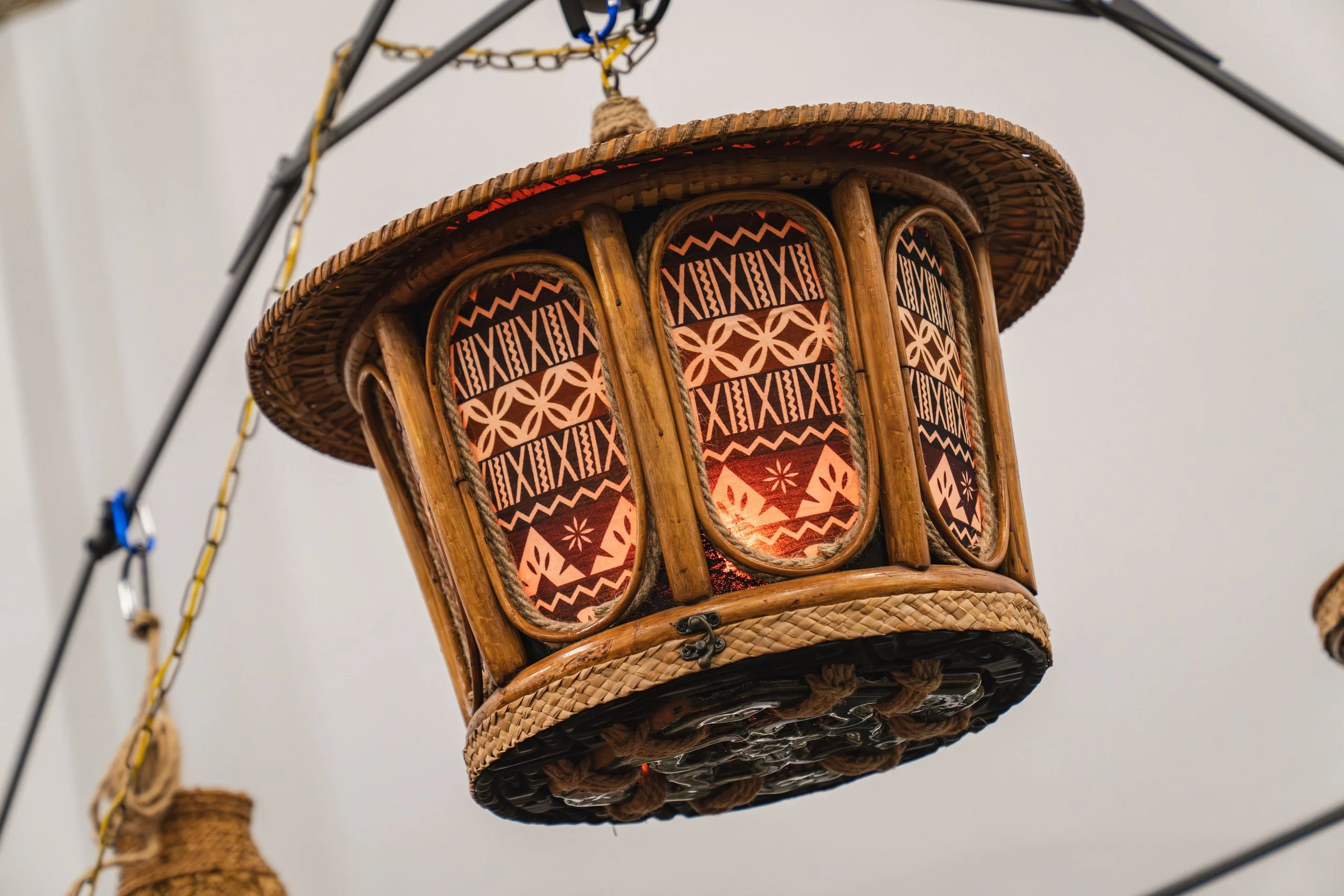 Close-up of a woven basket chair with tropical patterns on the seat, hanging from a chain and metal framework, against a plain background.