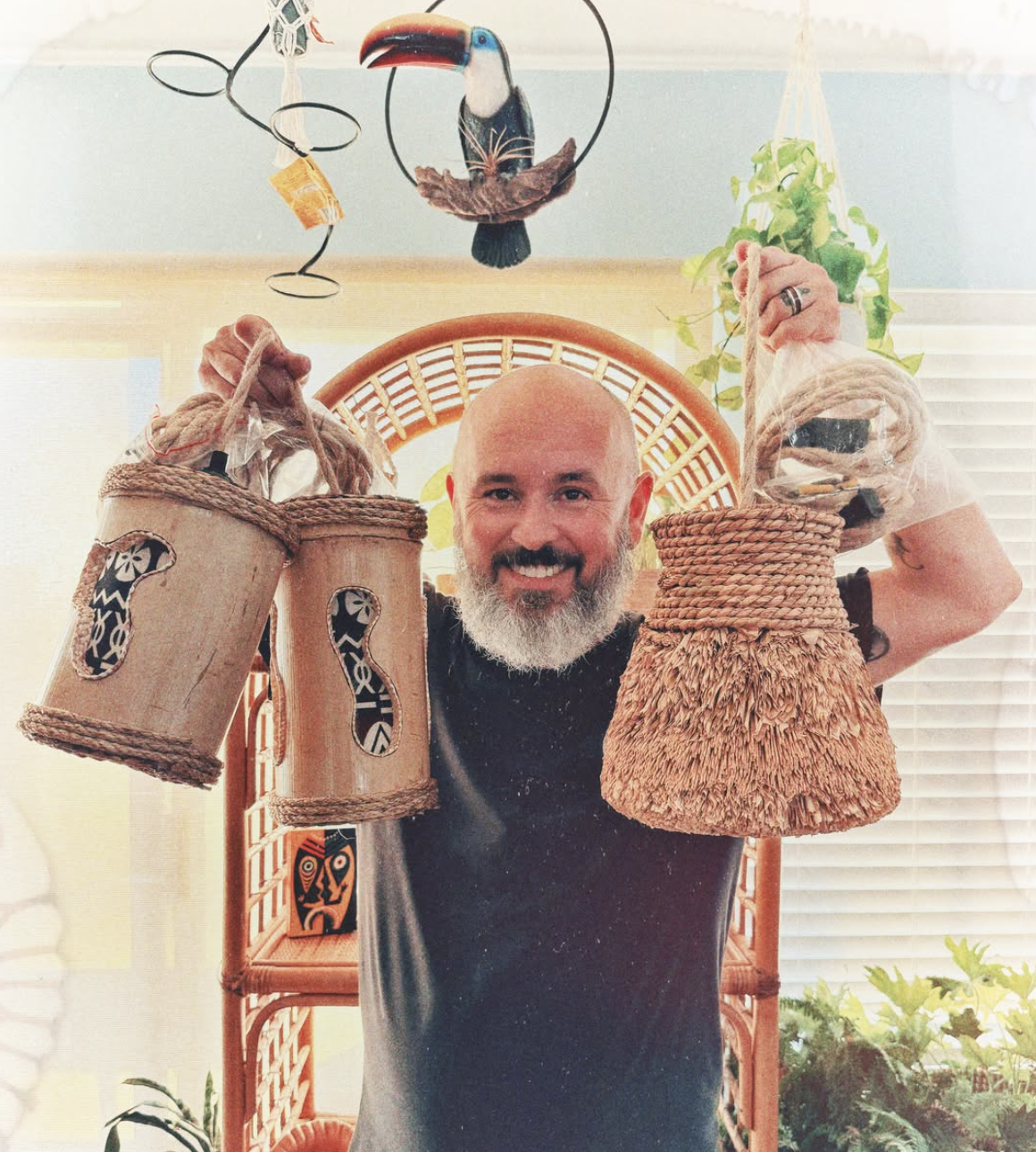A smiling man with a beard holds up four woven baskets, two in each hand, with a tropical-themed decor background including a hanging toucan and plants.