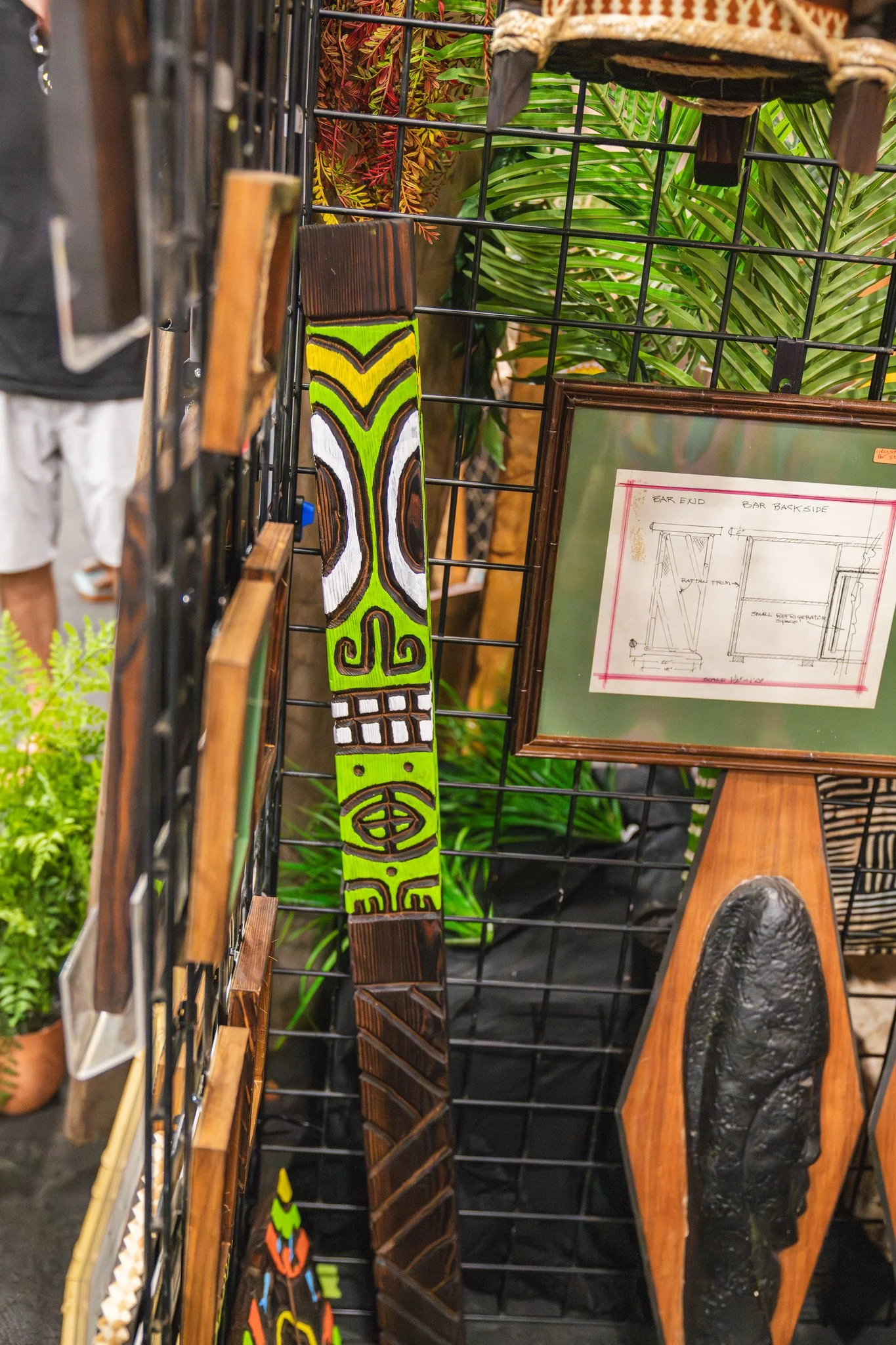 A display of decorative items on a metal grid, including a colorful painted wooden piece with abstract patterns, a framed drawing of furniture, a carved stone object, a wooden paddle, and lush green plants in the background.