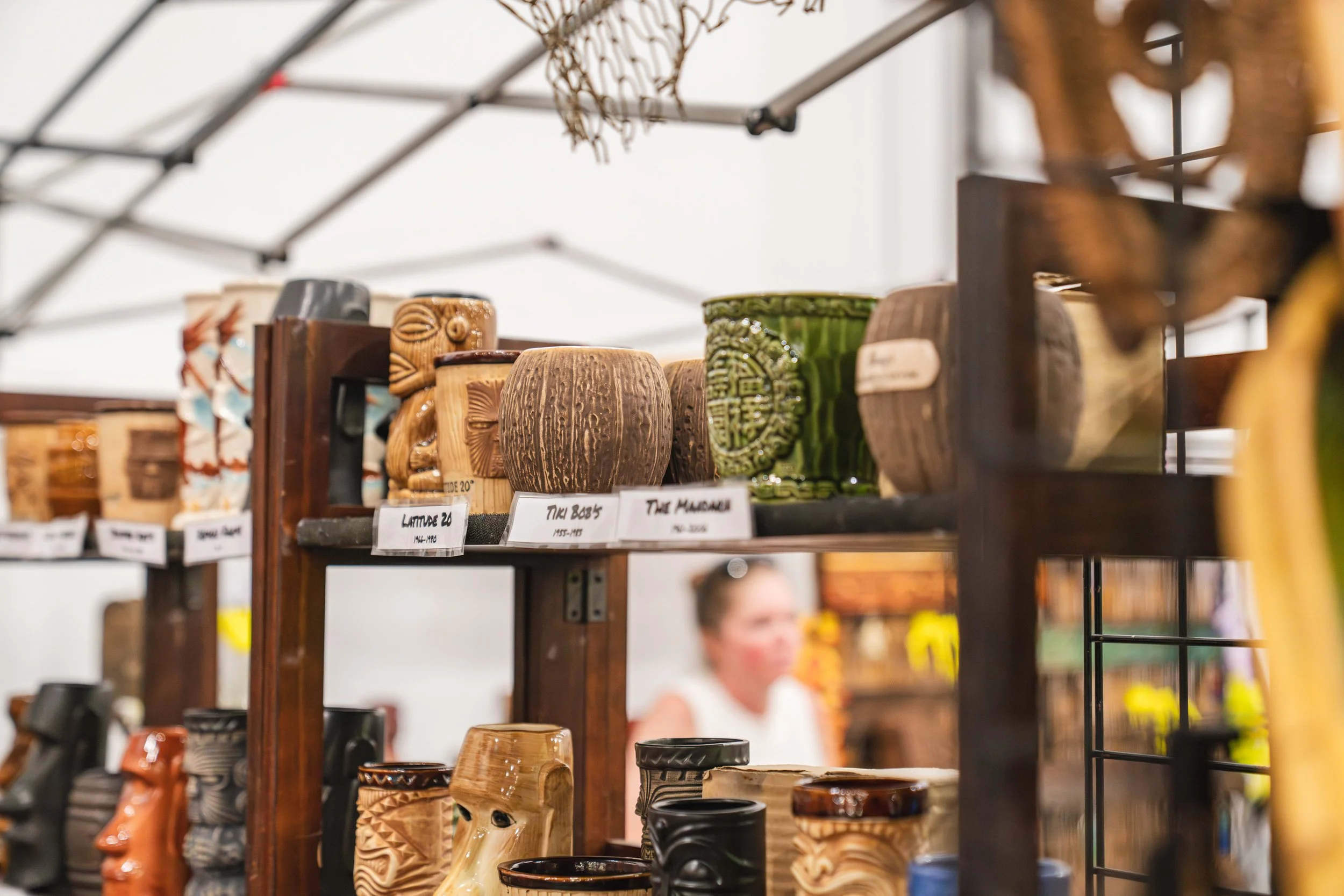 Various carved wooden and ceramic mugs displayed on shelves at a market, with a person blurred in the background.
