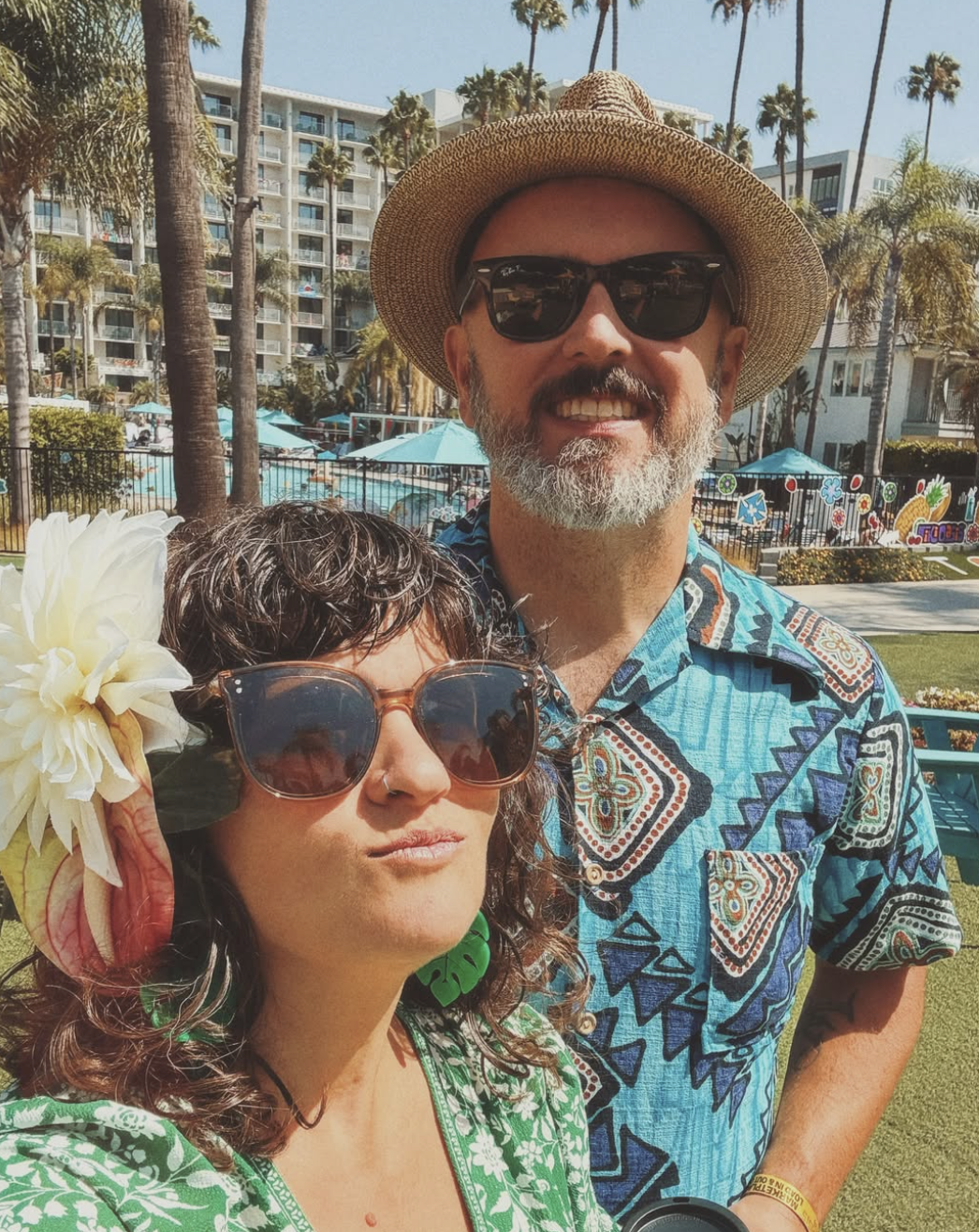 A man and woman taking a selfie outdoors at a sunny location, with palm trees, a pool, and a multi-story building in the background. They are wearing colorful, tropical-themed clothing and sunglasses, and appear to be enjoying a vacation or day out.
