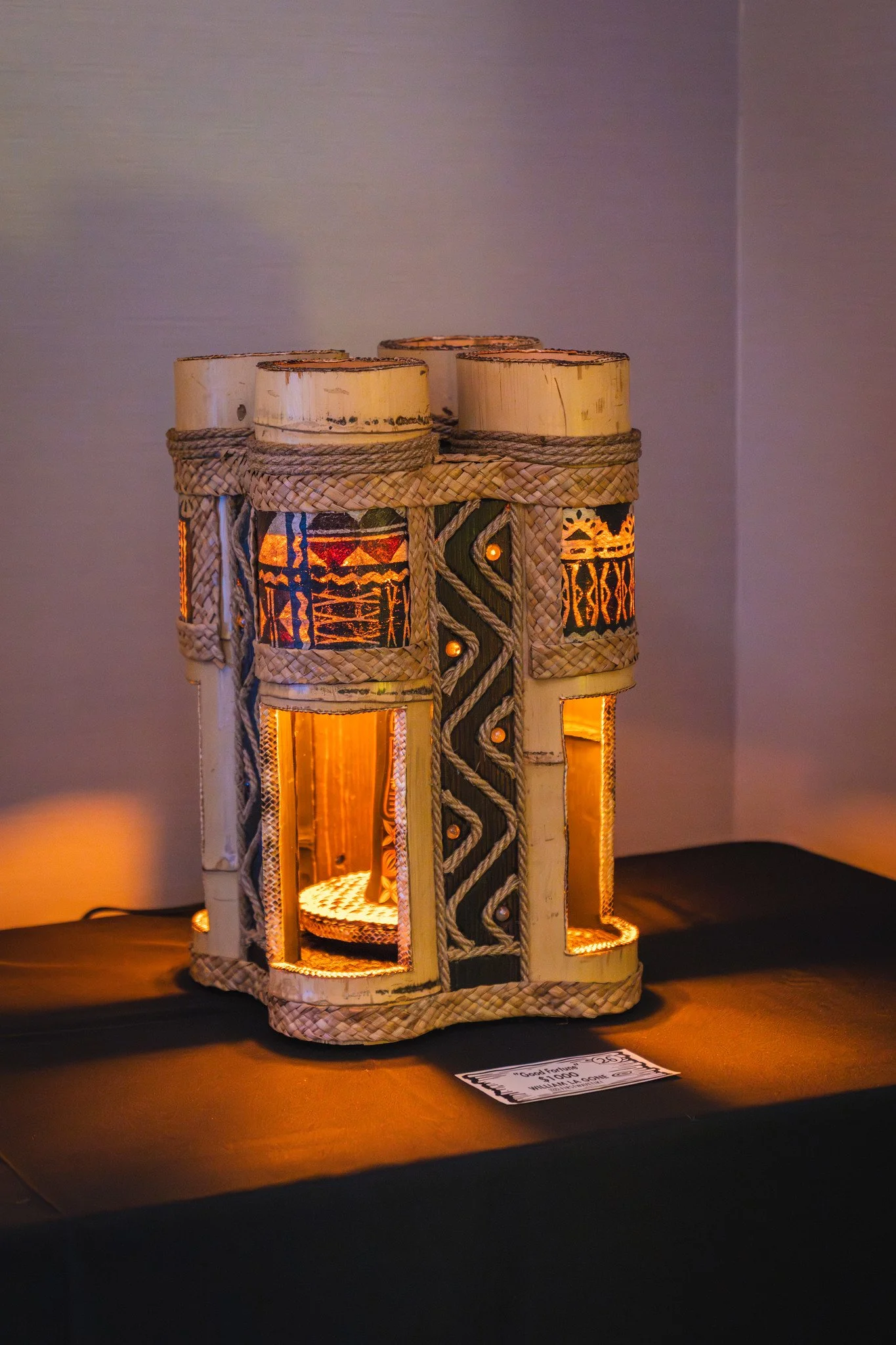 A decorative lamp made of woven bamboo and illuminated with internal lighting, displayed on a black surface with a small informational sign in front of it.