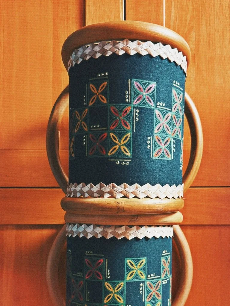 A decorative stool with a dark blue fabric cover embroidered with colorful geometric floral patterns, topped with a smooth wooden rim and supported by a wooden frame, reflected on a shiny surface.