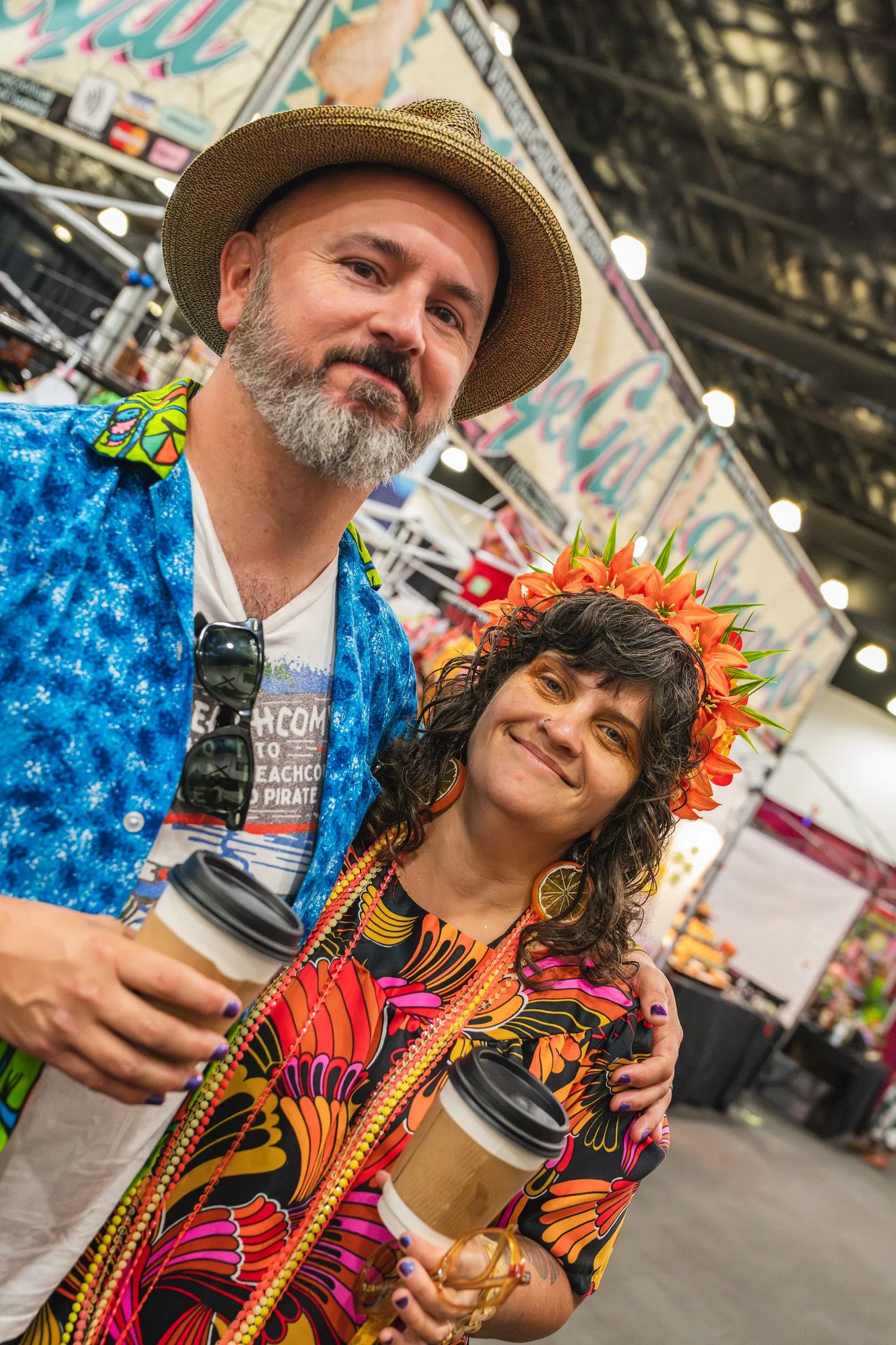 A man in a patterned shirt and straw hat and a woman in a tropical dress and flower crown are smiling and holding coffee cups at an indoor market or event.