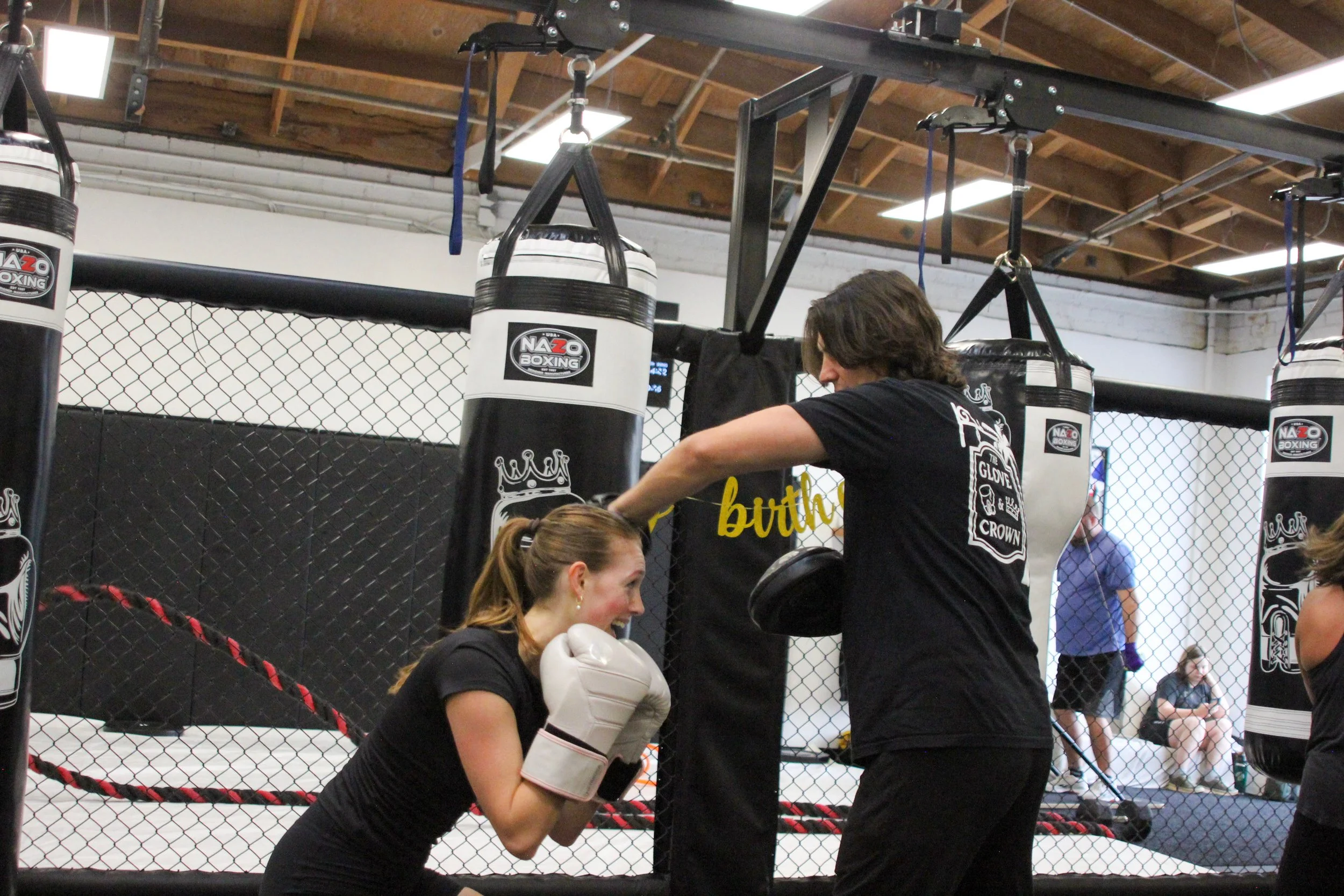 A woman practicing boxing with a trainer in a boxing gym, with punching bags hanging and people watching in the background.