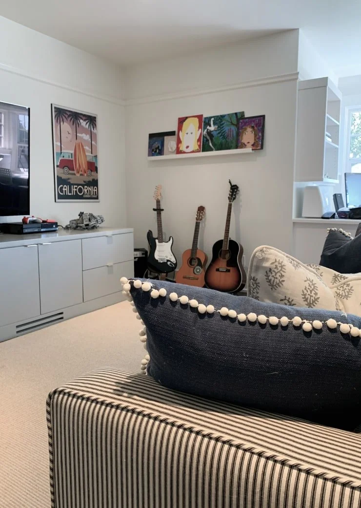 Living room corner with three guitars, a picture of a surfer in California and art on shelves, a television, and striped and solid pillows on a sofa.