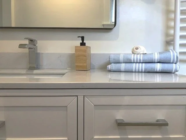 Bathroom sink counter with a silver faucet, a wooden soap dispenser, neatly folded blue and white towels, and a seashell on top. A mirror and a small part of a heating radiator are also visible.