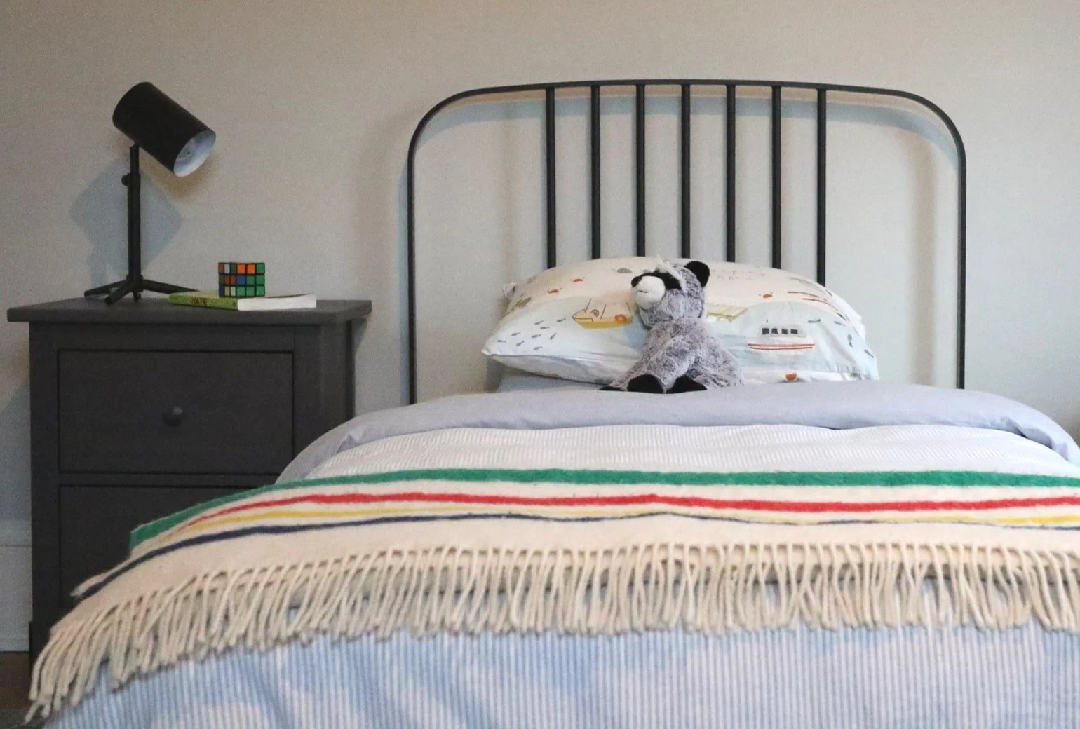 A neatly made bed with a grey blanket, topped with a striped blanket with fringes, and a pillow with a cartoon boat design. A stuffed animal resembling a raccoon sits on the pillow. To the side, there is a grey nightstand with a black desk lamp, a small colorful cube puzzle, and a closed book.