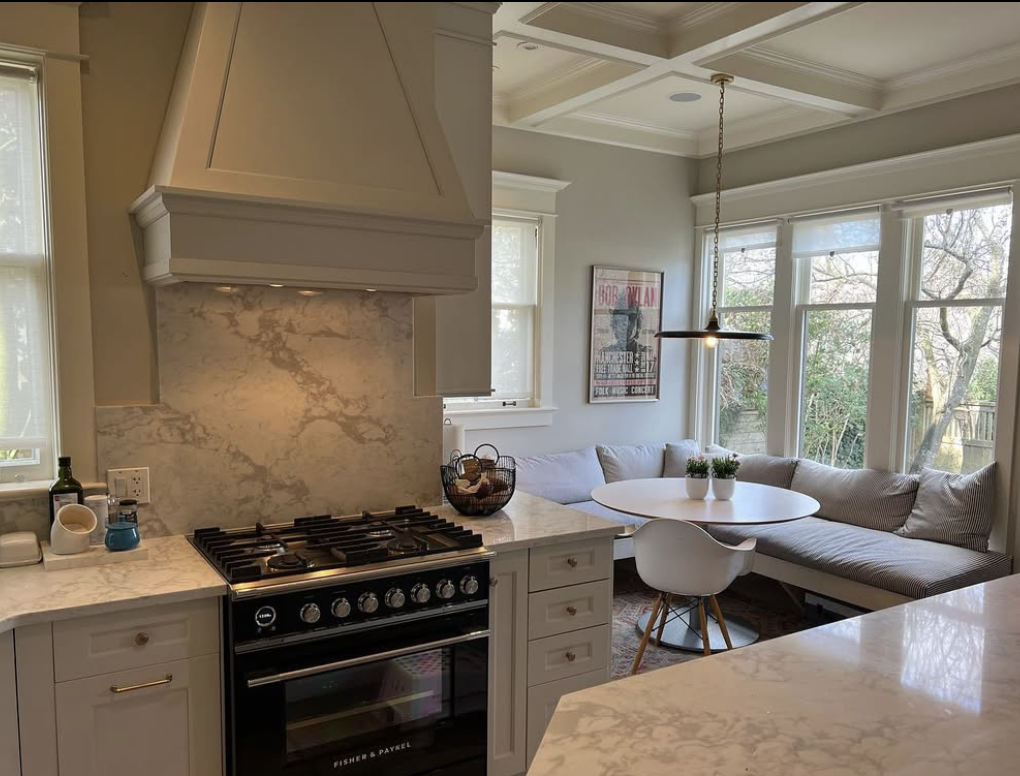 Kitchen with stovetop, marble backsplash, and a seating area with a white round table, a white chair, and a built-in bench with pillows near large windows.