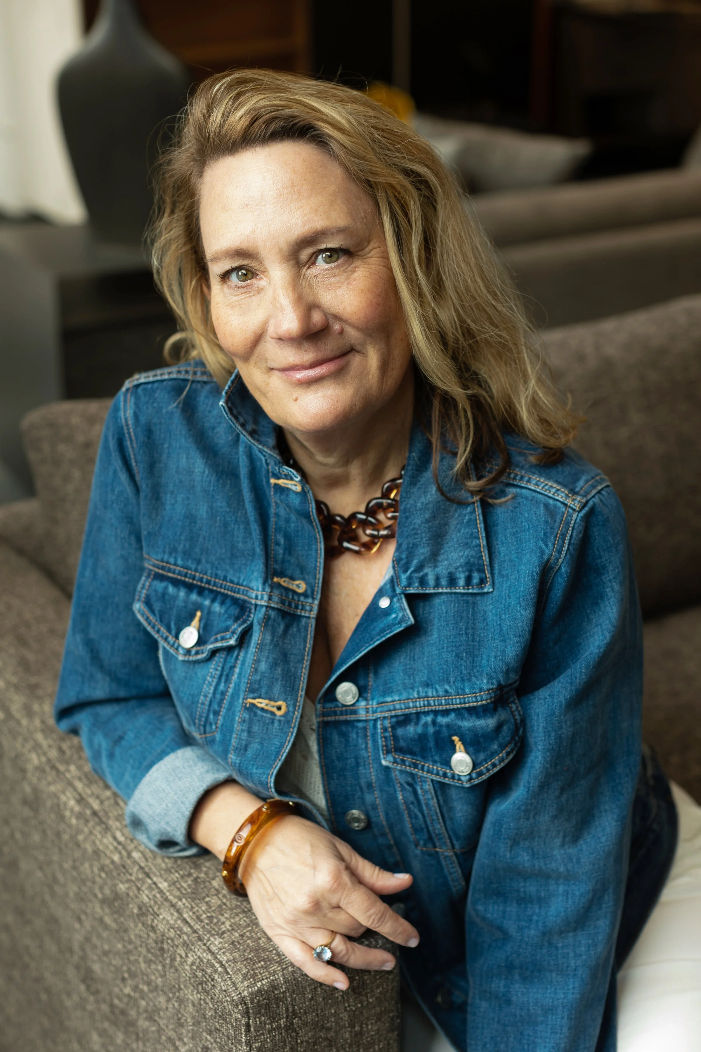 A woman with blonde hair sitting on a couch, wearing a denim jacket and jewelry, smiling at the camera in a cozy indoor setting.