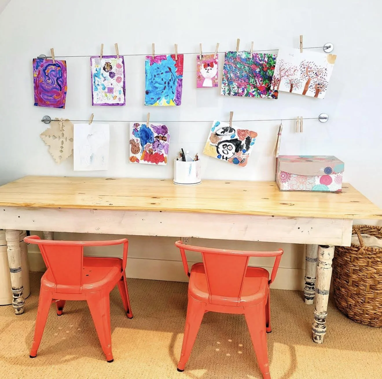 Children's art displayed on a string with clothespins above a wooden table with red chairs, art supplies in a container, and a decorative box, in a bright room.
