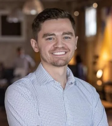 A smiling young man with short dark hair and a beard, wearing a light blue checkered shirt in an indoor setting with warm lighting and blurred background.