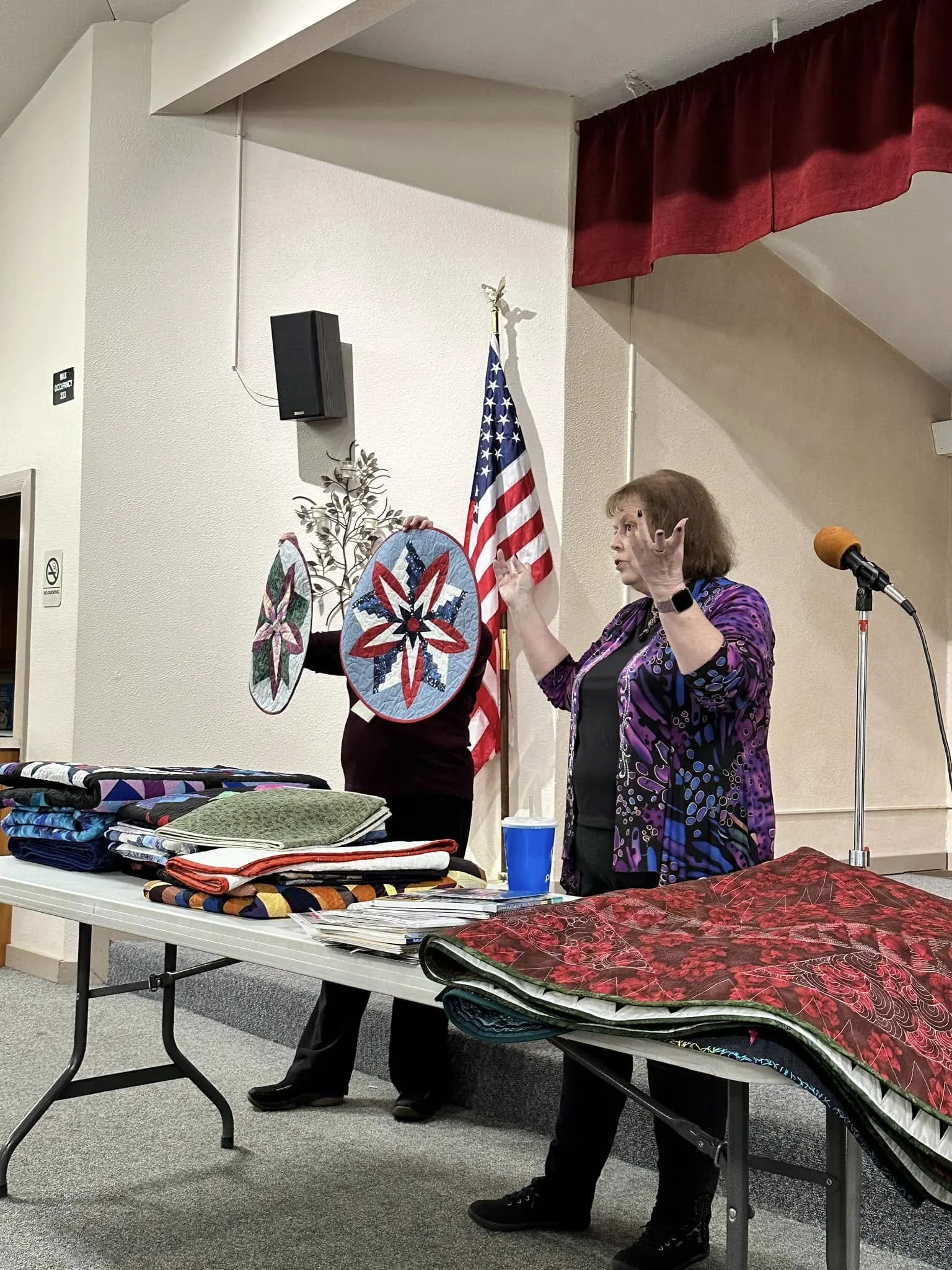 A woman stands at the front of the room demonstrating quilt techniques.