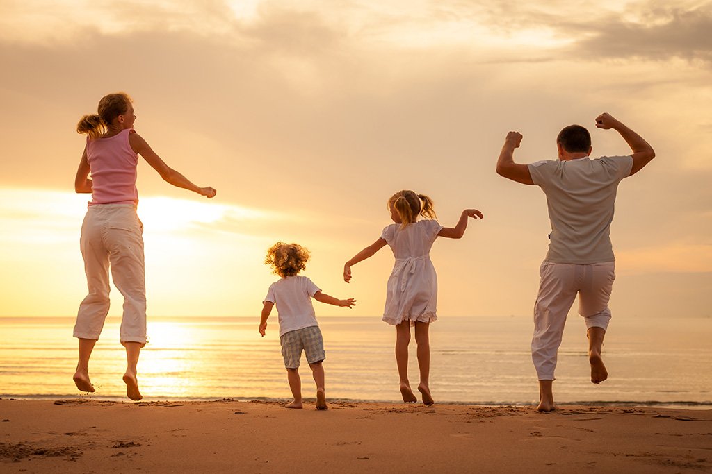 A family of four jumping on a beach at sunset, with the ocean in the background.