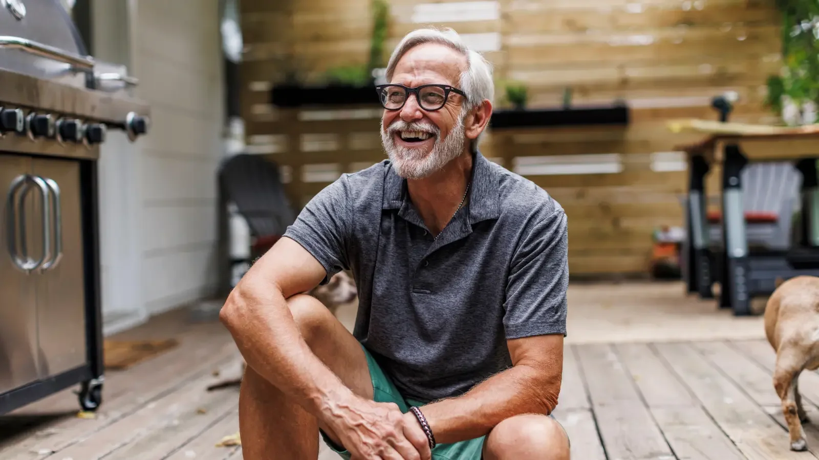 A smiling elderly man with glasses and a beard, wearing a gray polo shirt and green shorts, sitting on a wooden deck outdoors.