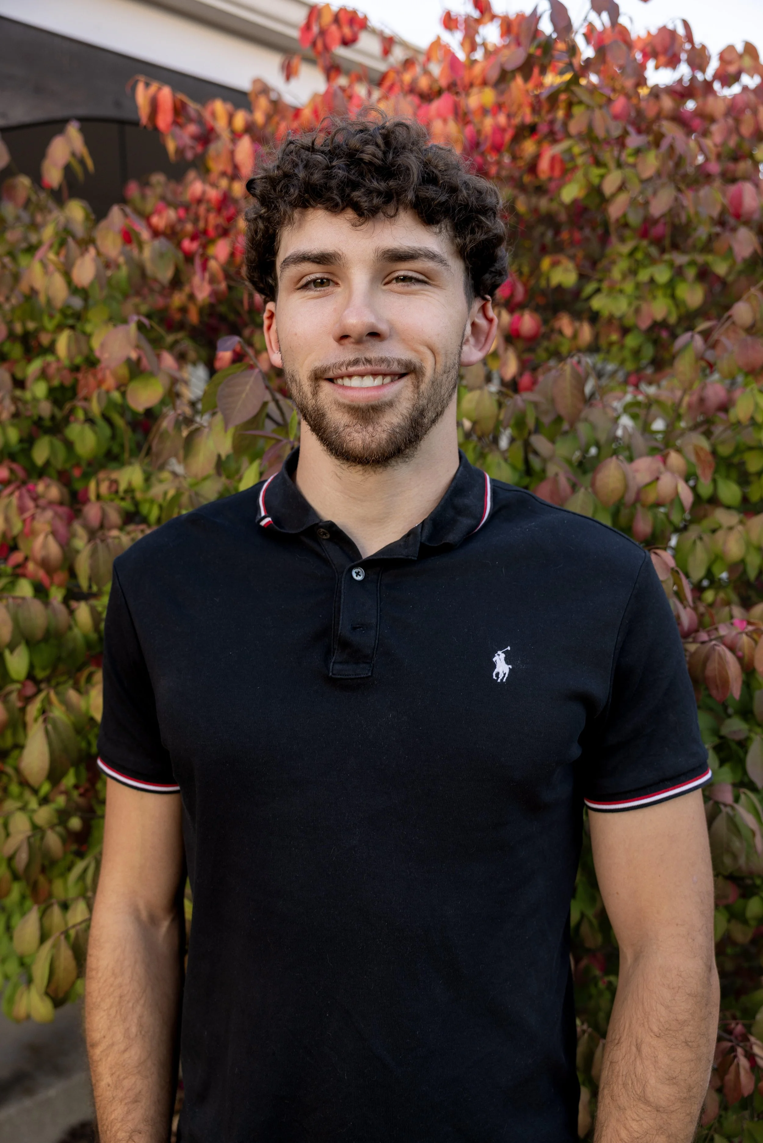 A young man with curly brown hair and a beard, smiling, wearing a black polo shirt with a small white logo, standing outdoors in front of a bush with reddish and green leaves.