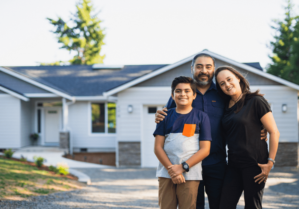 A family of three standing in front of a house, smiling and hugging each other on a sunny day.