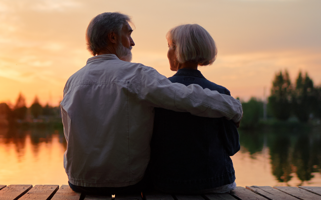 An elderly couple sitting close together on a dock by the water during sunset.