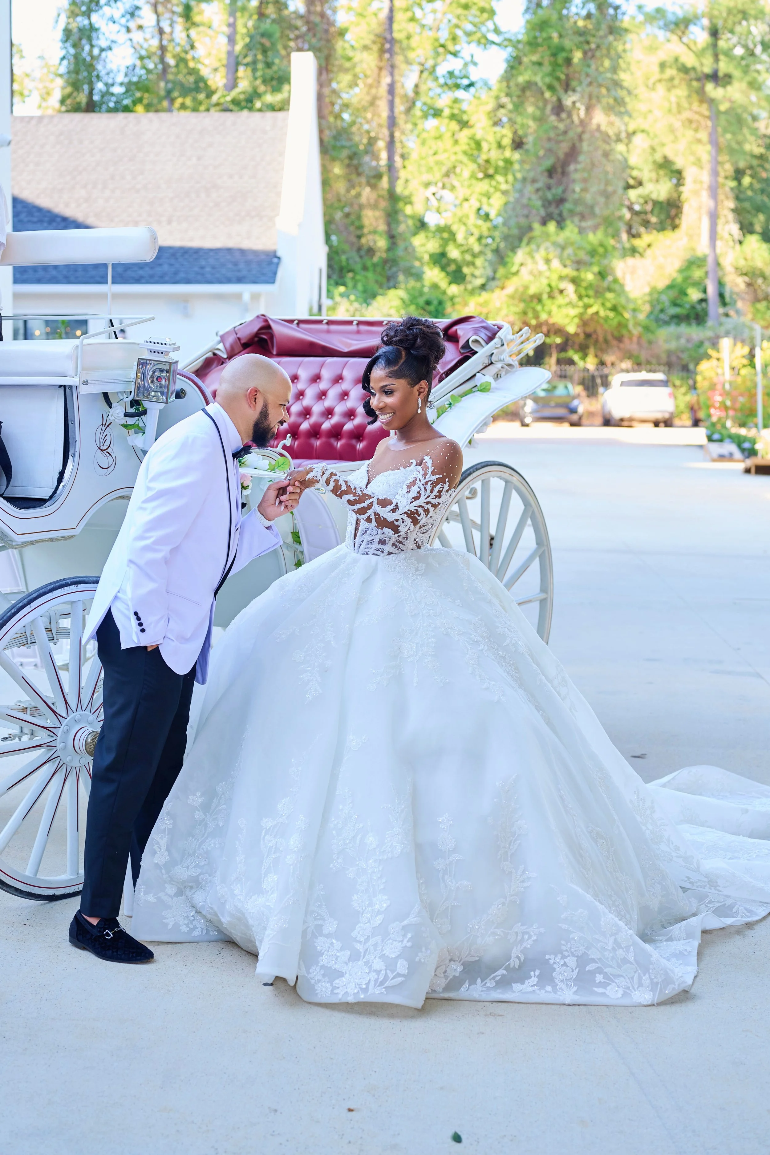 A bride in a white wedding gown and a groom in a white suit with black pants are smiling and holding hands in front of a white horse-drawn carriage with a red interior, outdoors during daytime.