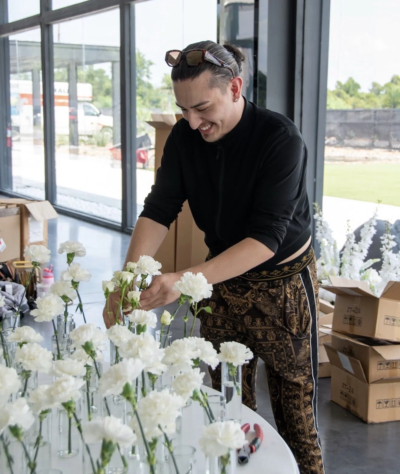 A person arranging white carnations in vases inside a brightly lit room with large windows.