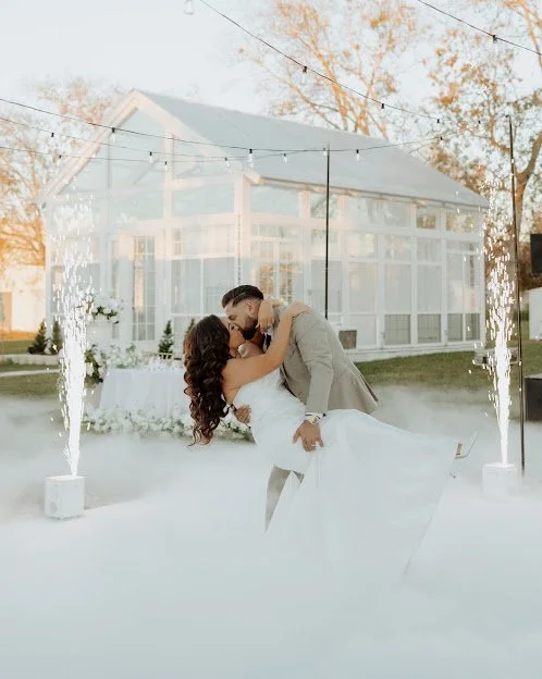 A newlywed couple sharing a kiss during their wedding reception. The bride is wearing a white gown, and the groom is in a beige suit. They are dancing outdoors in front of a glass house, with sparklers and string lights creating a festive atmosphere.