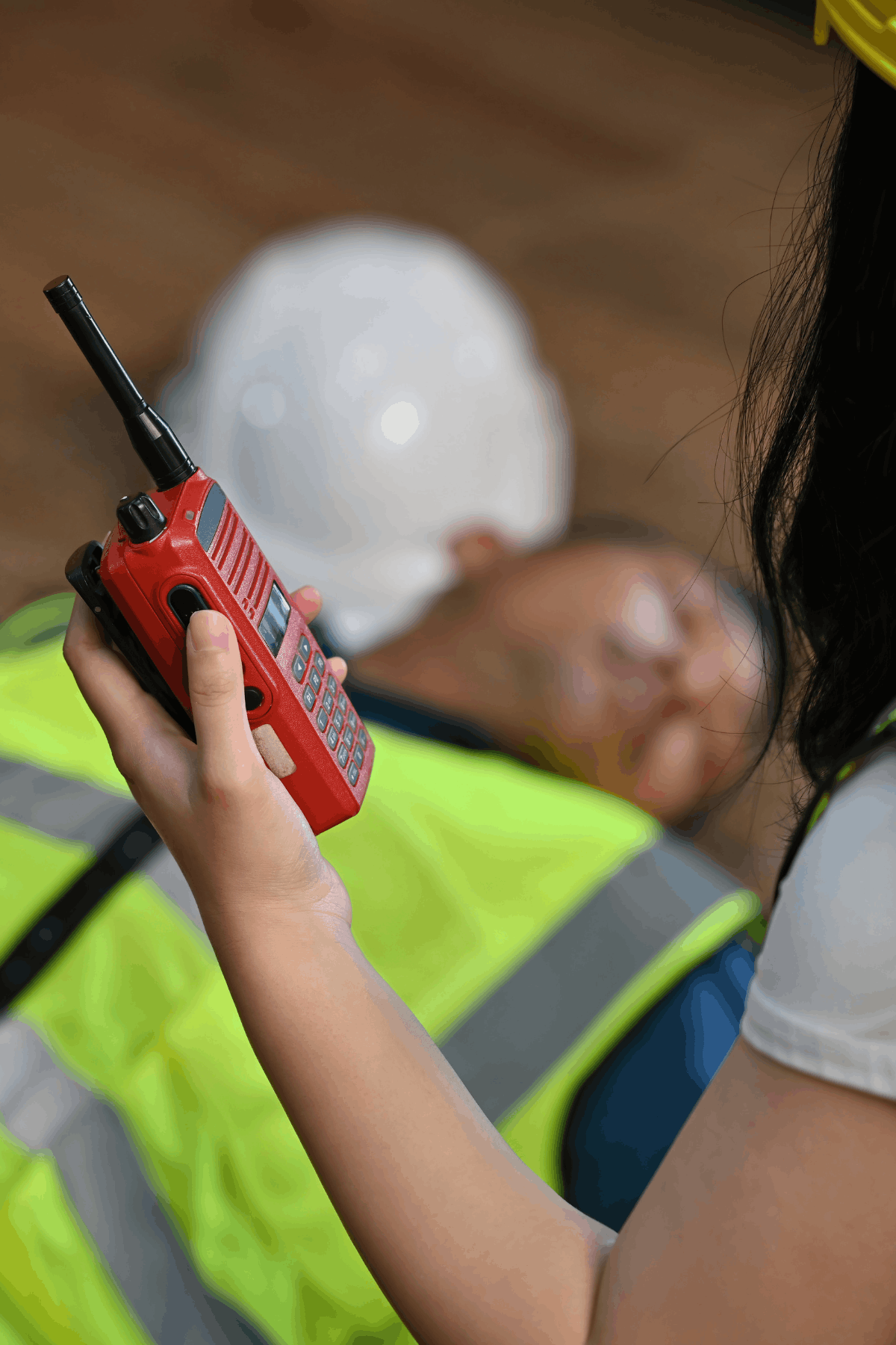 A person in a yellow hard hat and safety vest holds a red walkie-talkie, with another person in a white hard hat and safety vest lying on the ground in the background.
