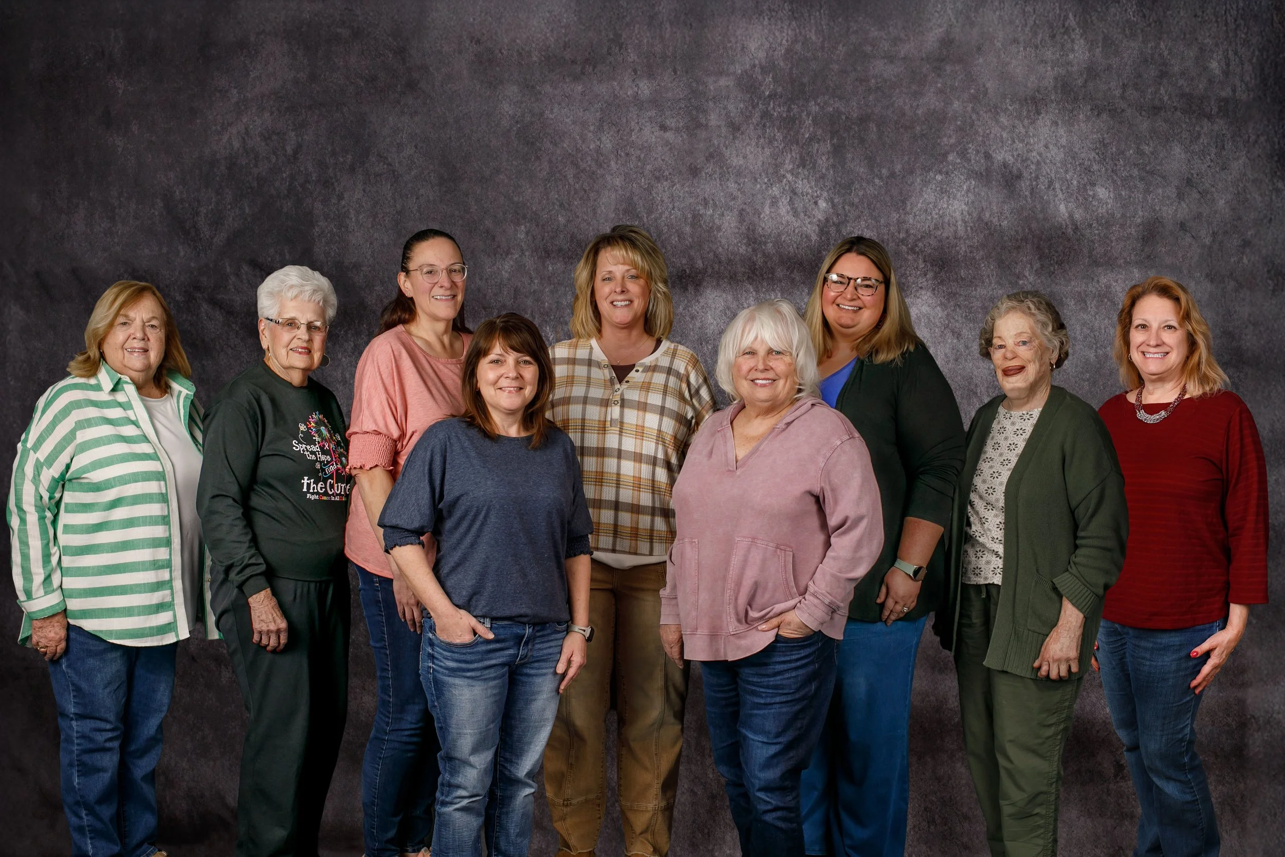 A group of eleven women of various ages standing together in front of a dark gray textured background, smiling at the camera.