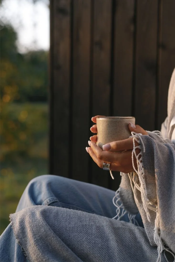 Person sitting outdoors holding a ceramic mug, wearing a beige sweater with frayed edges and blue jeans, with a dark wooden wall behind them and a blurred natural background.