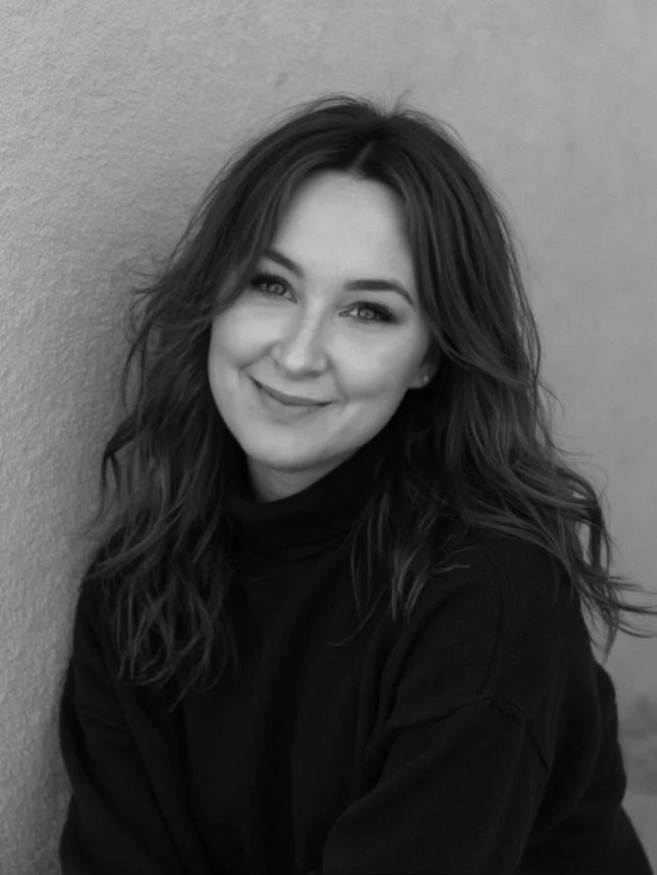 Black and white portrait of a woman with wavy hair smiling at the camera, wearing a black turtleneck, against a plain wall background.