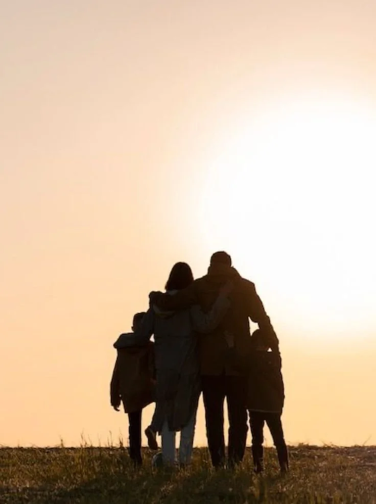A group of people standing outdoors on grass, silhouetted against a bright setting sun, with some embracing each other.
