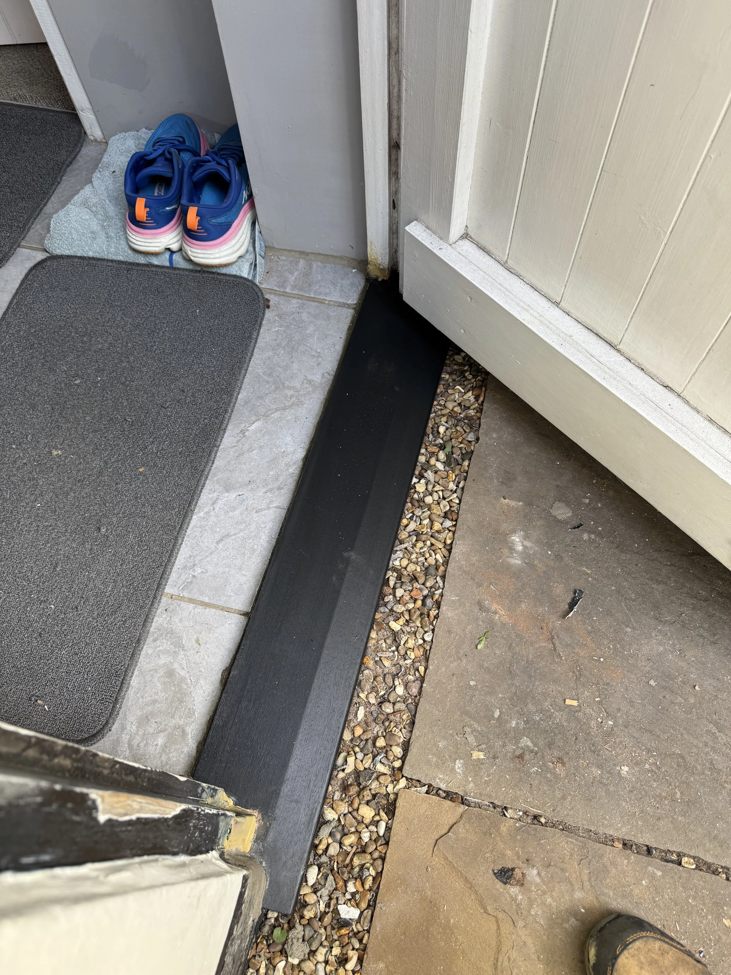 Entryway with shoes, rugs, tiles, a black threshold, gravel, and concrete flooring.