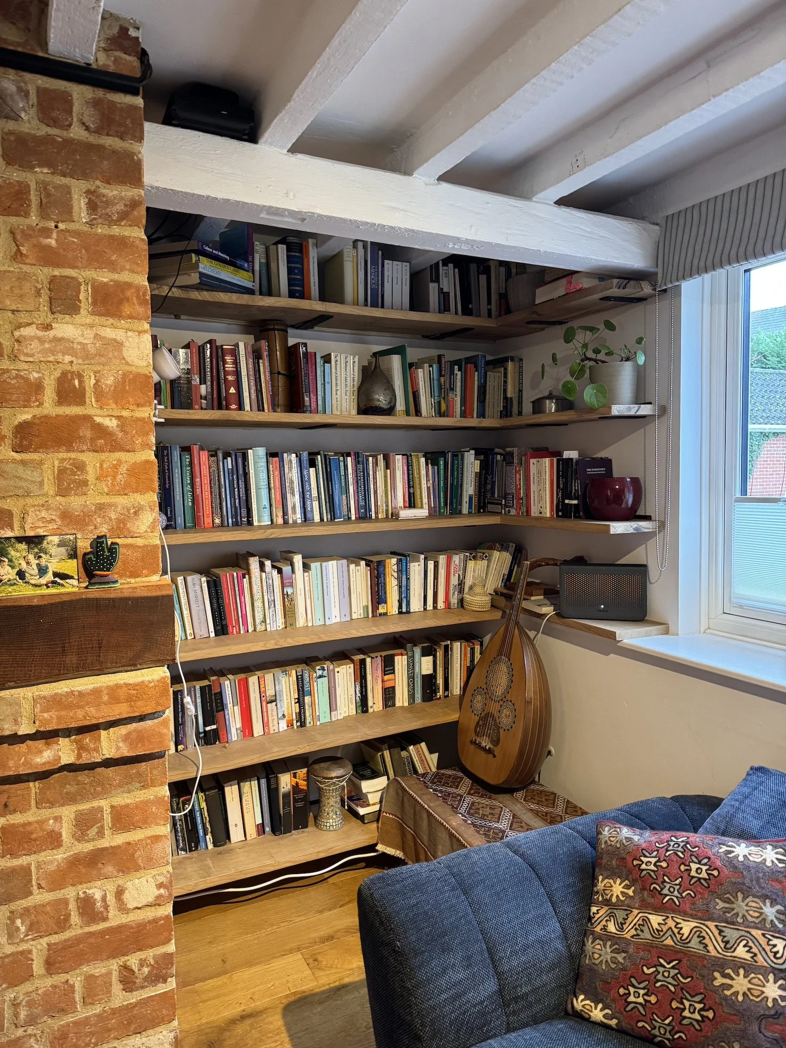Bookcase filled with books next to a window, with various decorative items including a potted plant, a wine glass, and a stringed musical instrument, in a cozy living room with exposed brick wall and wooden flooring.